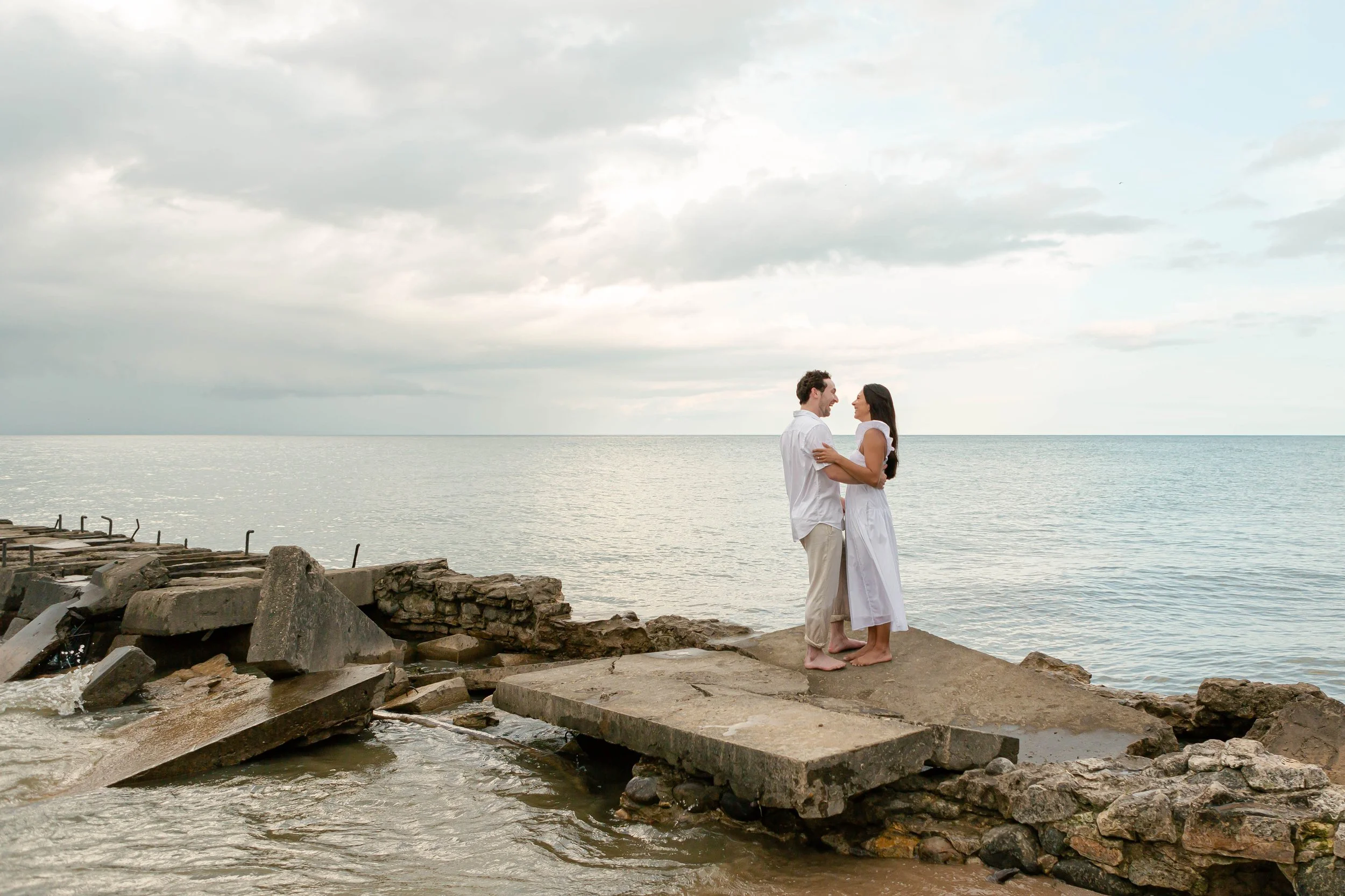 A couple in white clothing standing close together on a rocky jetty by the water, looking at each other with the ocean and cloudy sky in the background.