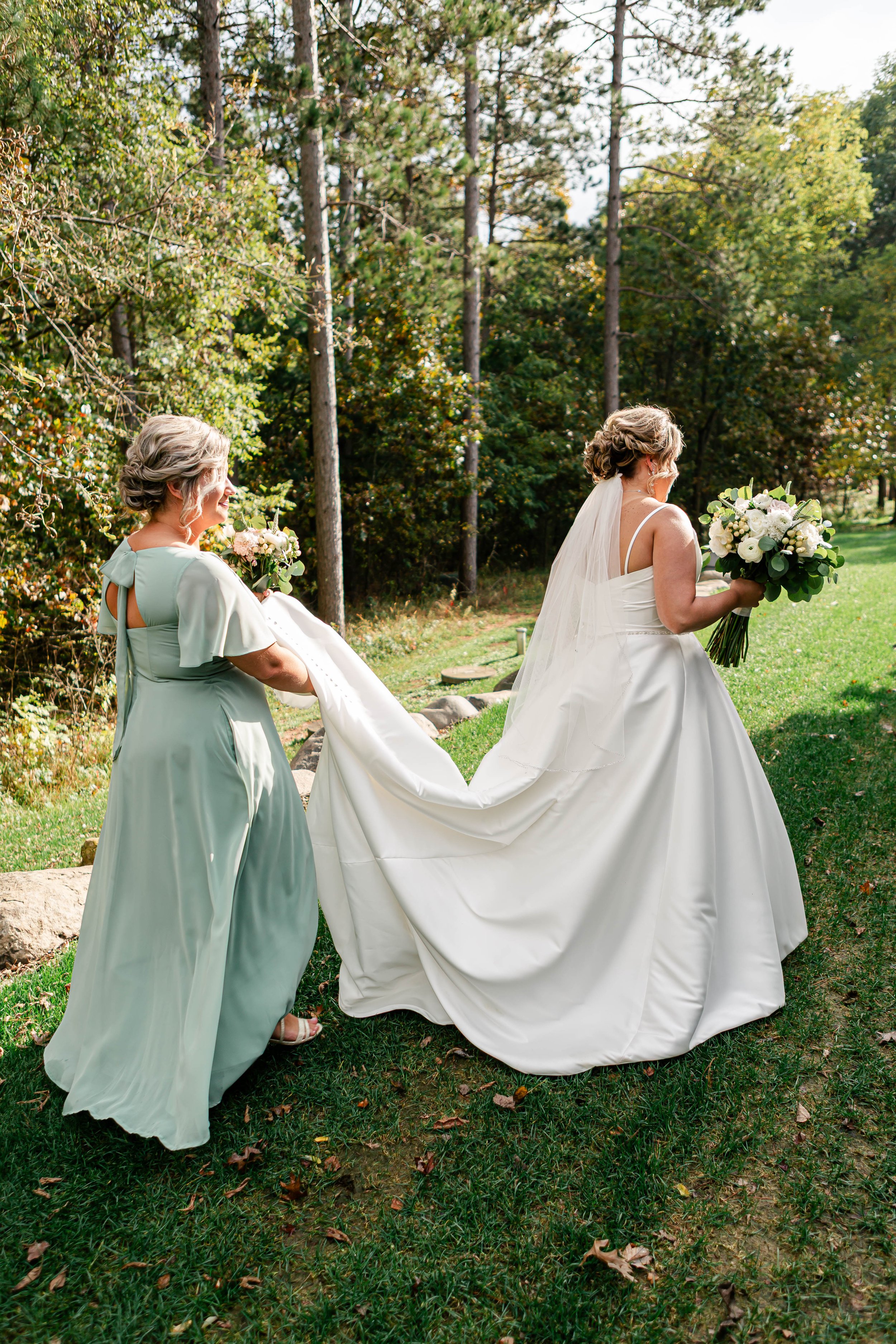 A bride in a white wedding gown holding a bouquet of white flowers walks through a green outdoor area with trees, while a woman in a light green dress holds the train of her dress.