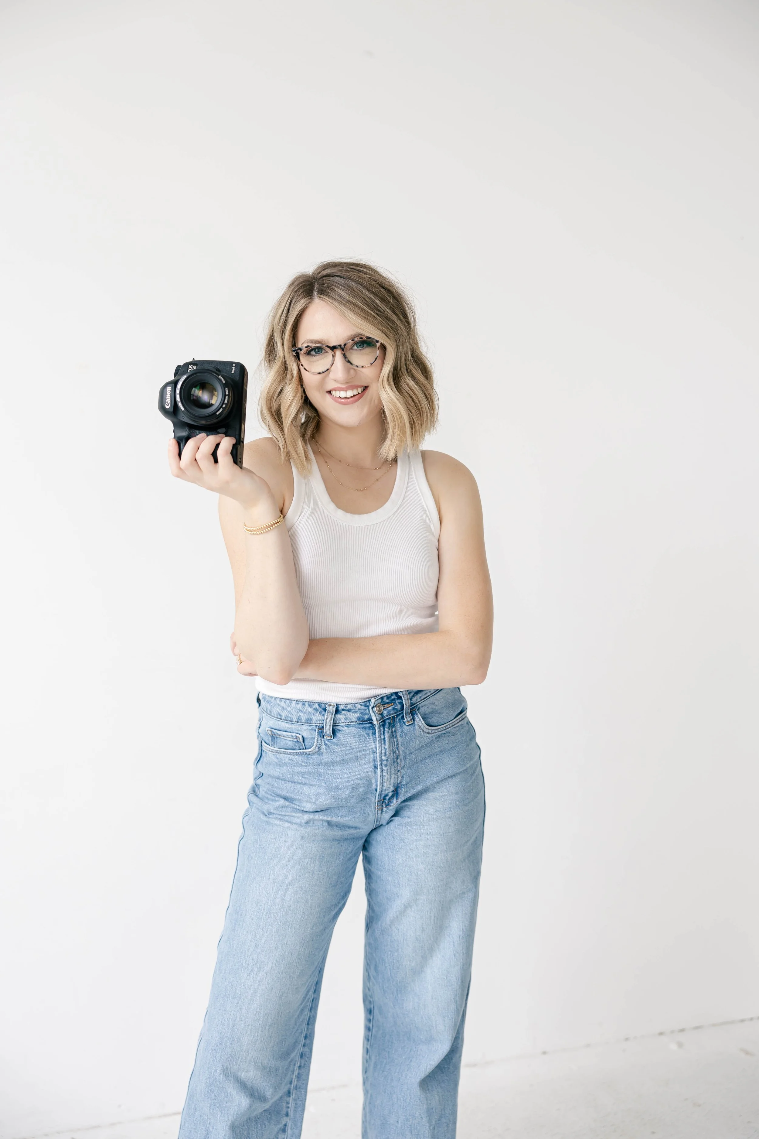 A woman with glasses and blonde hair smiling and holding a camera in front of a white background.