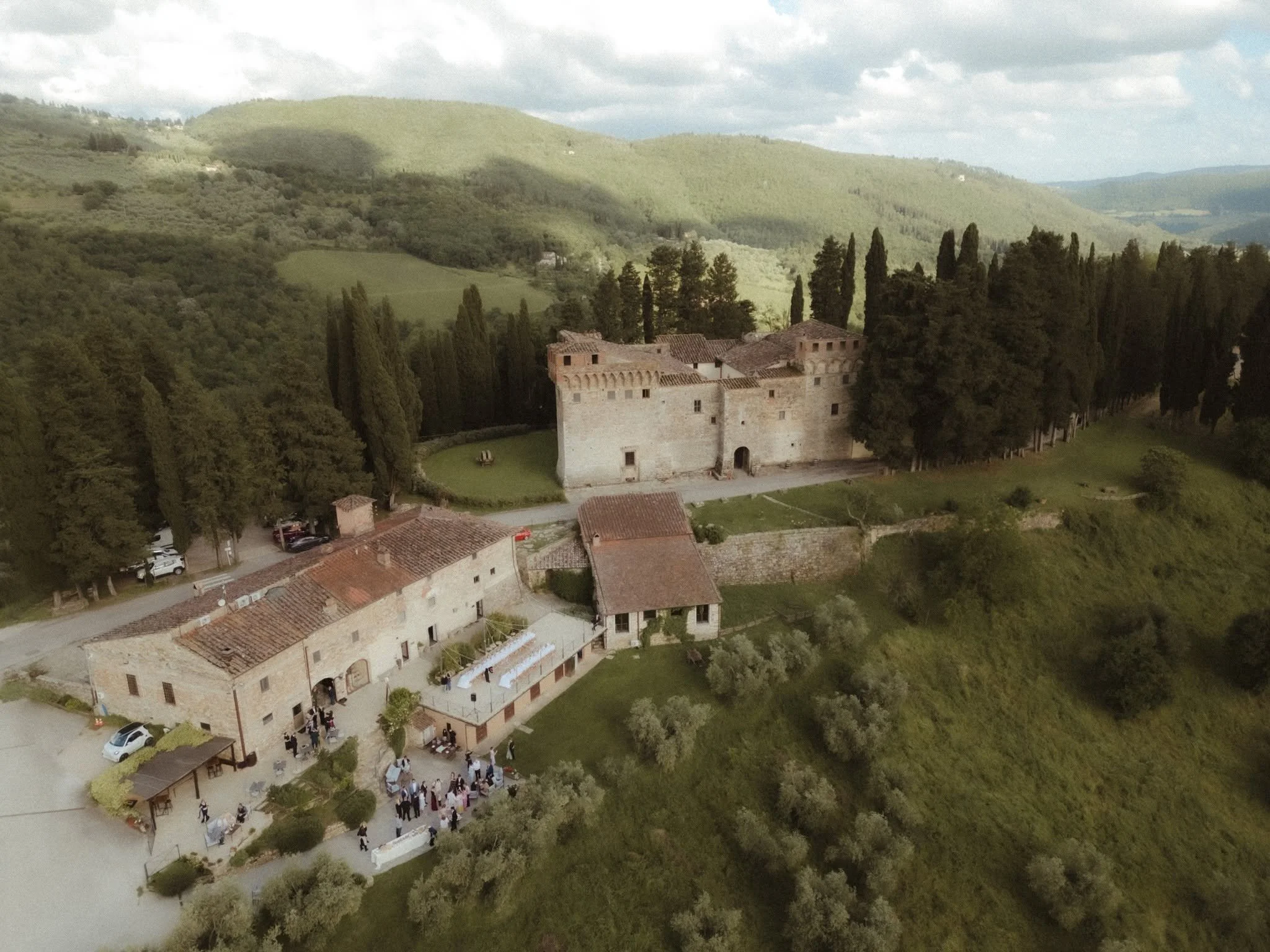 Aerial view of a historic stone castle with red-tiled roofs, surrounded by tall cypress trees, in a green hilly landscape with a cloudy sky.