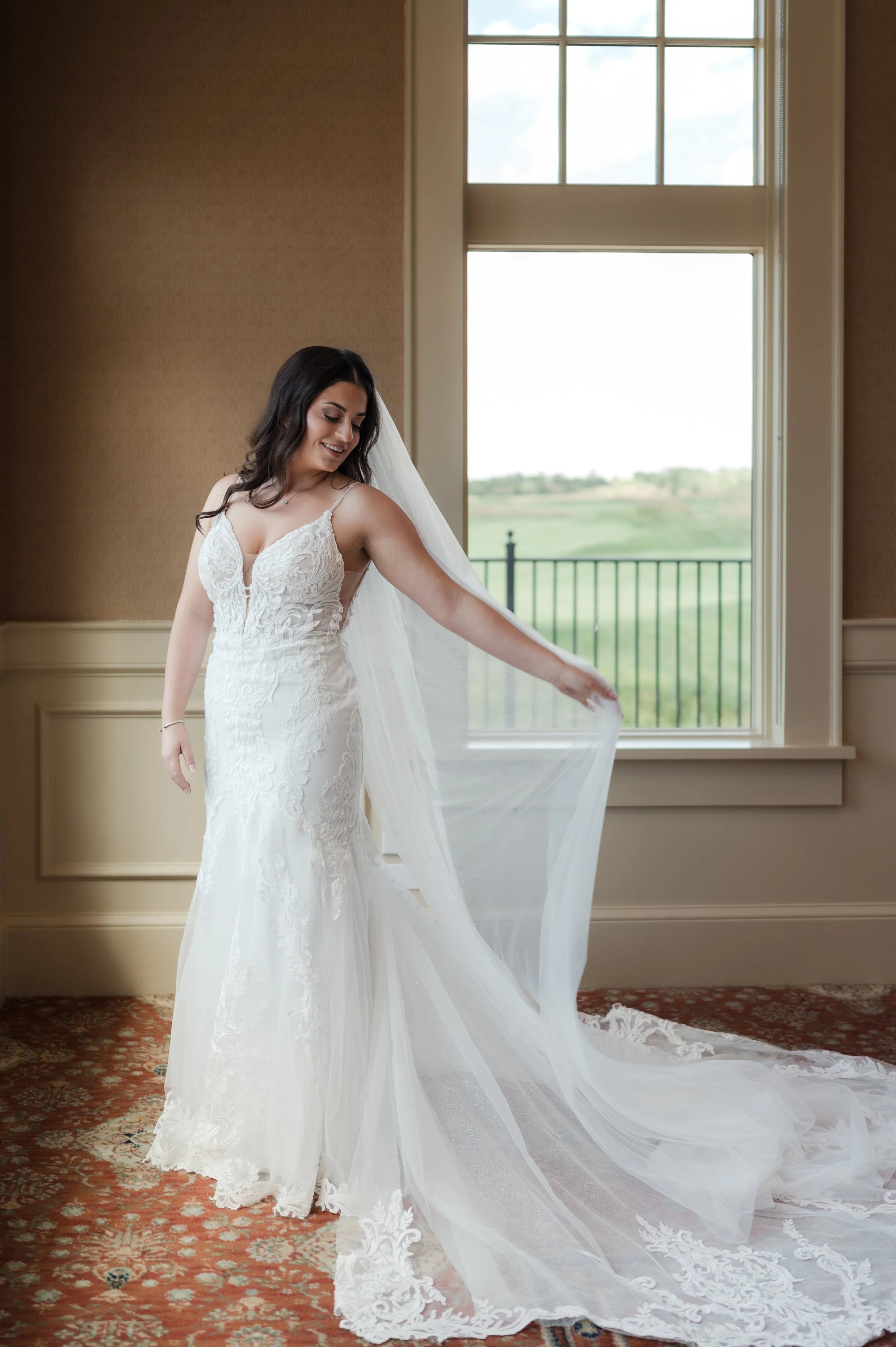 A bride in a white lace wedding dress standing indoors, holding her veil, with large windows showing a green landscape outside.