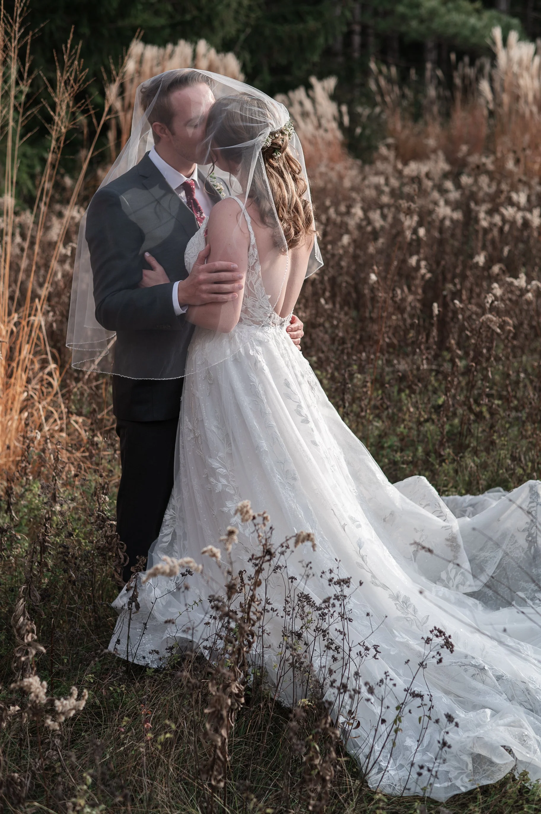 A bride and groom share a kiss outdoors, with the bride wearing a long white wedding gown and veil, and the groom in a dark suit, surrounded by tall grasses and trees.