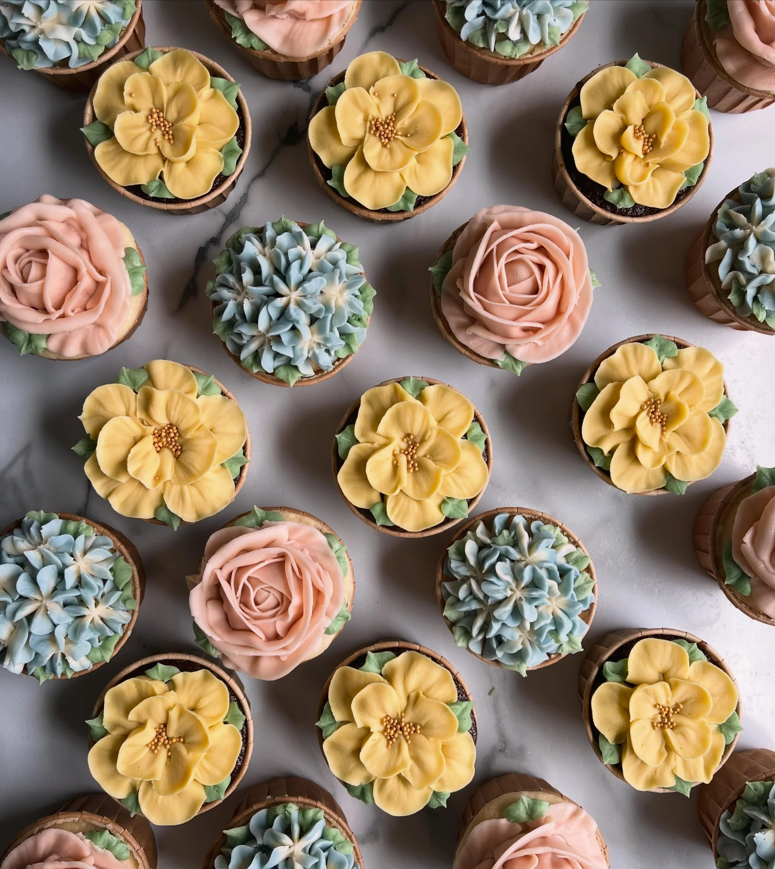 Assorted cupcakes decorated with pastel-colored frosting in floral and rosette shapes, arranged on a white marble surface.