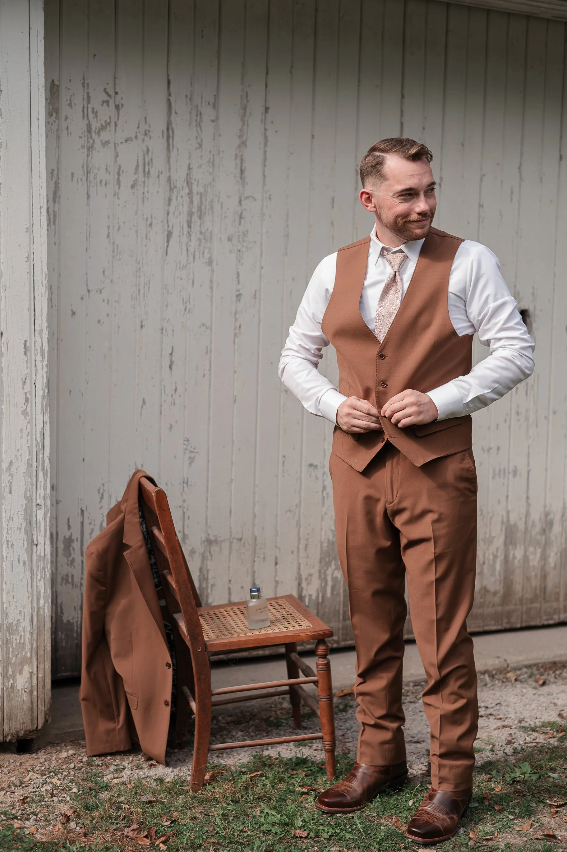A man in a brown vest and pants, white shirt, and tie standing outside in front of a weathered wooden wall, adjusting his vest. A wooden chair with a jacket draped over it and a small perfume bottle on its seat is nearby.