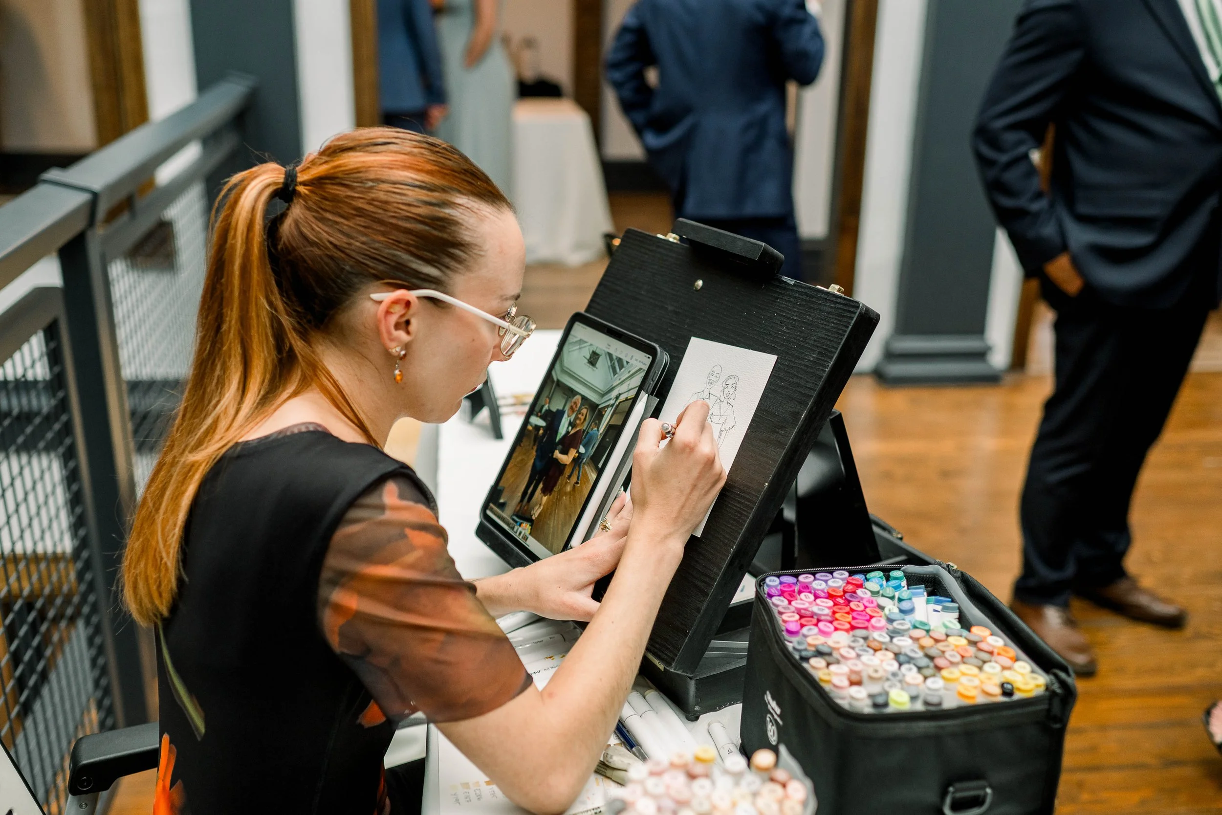 A woman with long red hair, glasses, and earrings, is seated at a table drawing on a tablet or notepad. She is wearing a black and orange top. In front of her are colorful markers in a case. Other people are visible in the background, standing and ta
