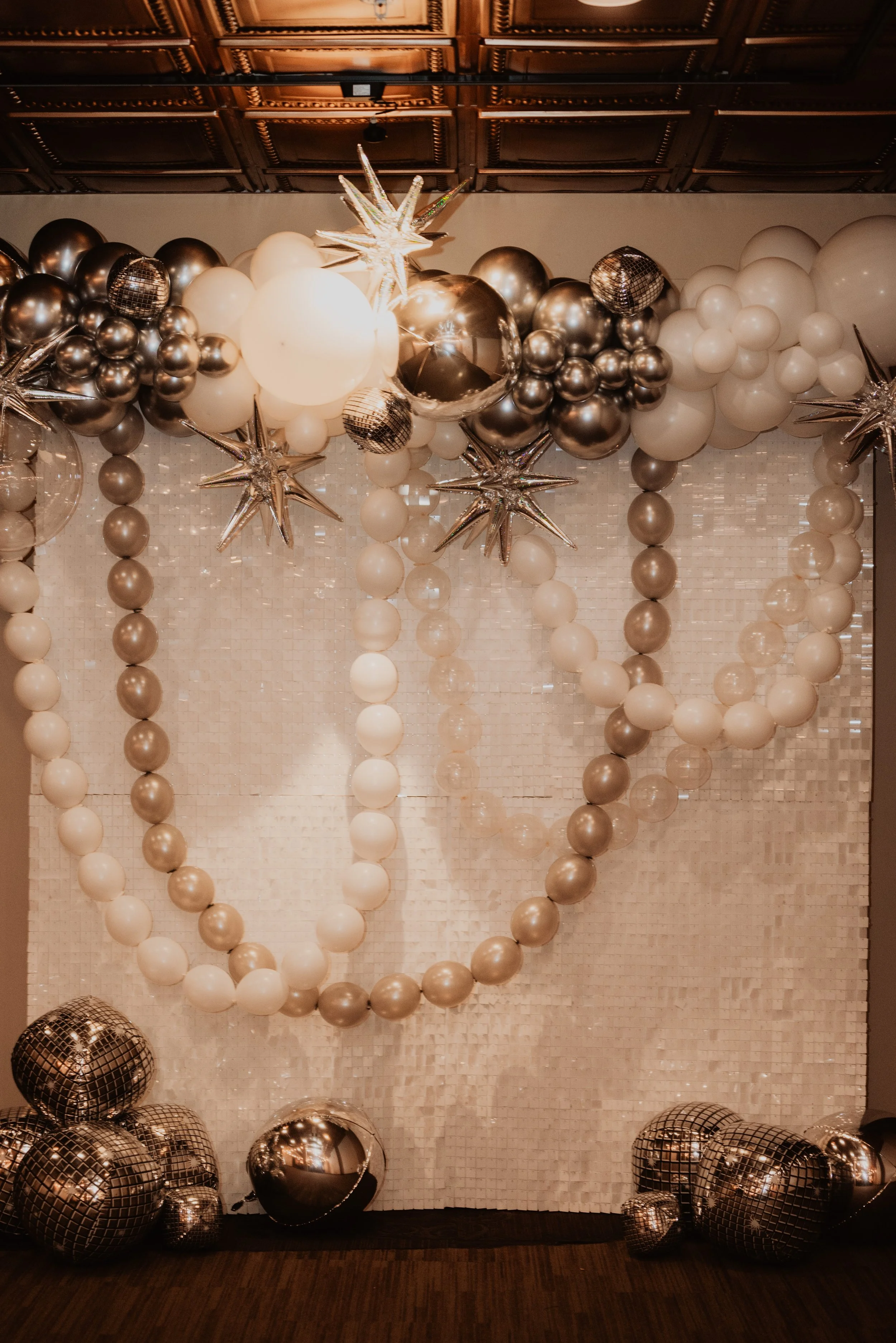 Balloon decoration with white, black, and silver balloons forming a large heart shape, with metallic star and disco ball balloons, against a textured white wall and decorative ceiling.
