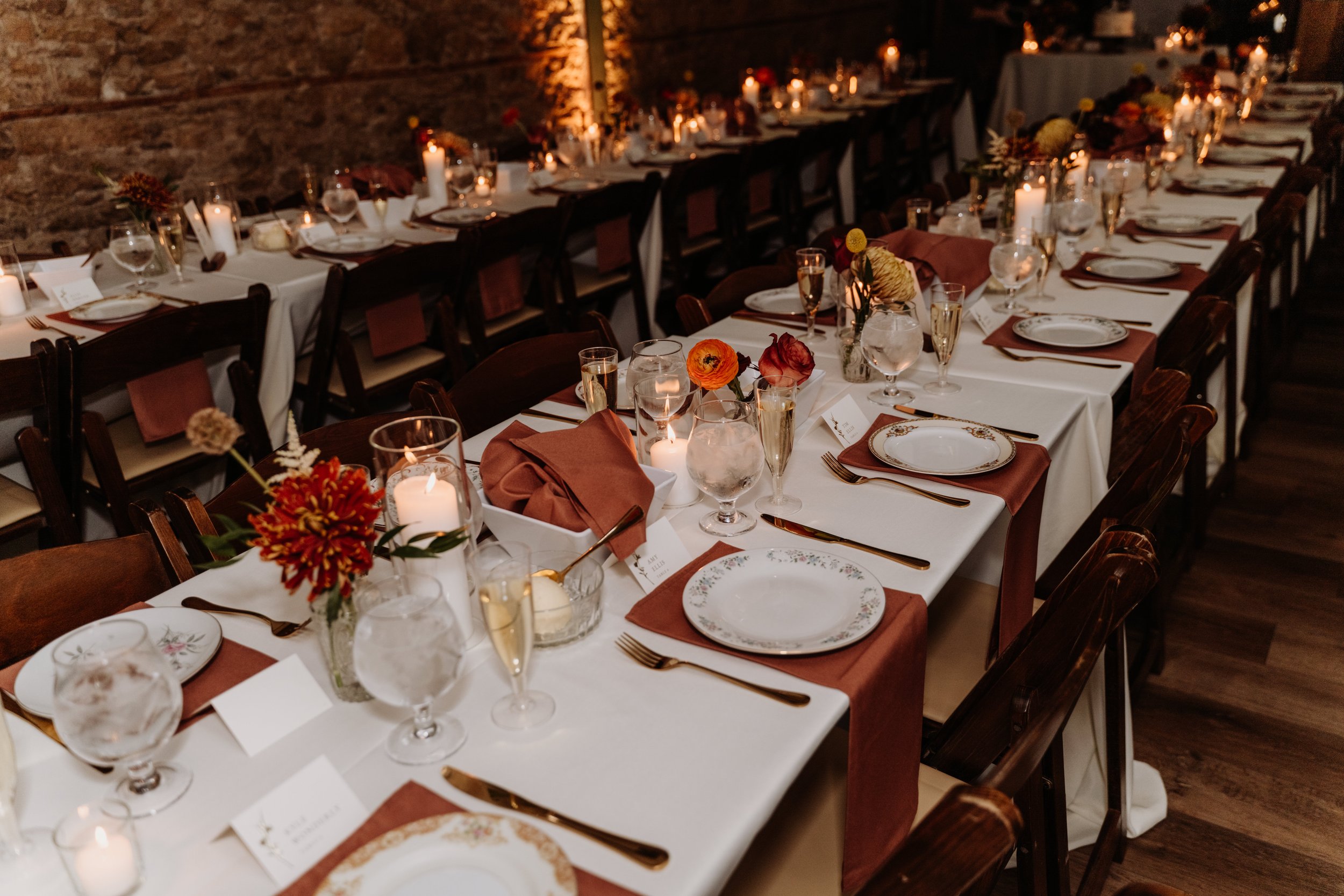 A long dining table set for an event with white tablecloths, floral centerpieces, candles, water glasses, and gold utensils, against a brick wall.