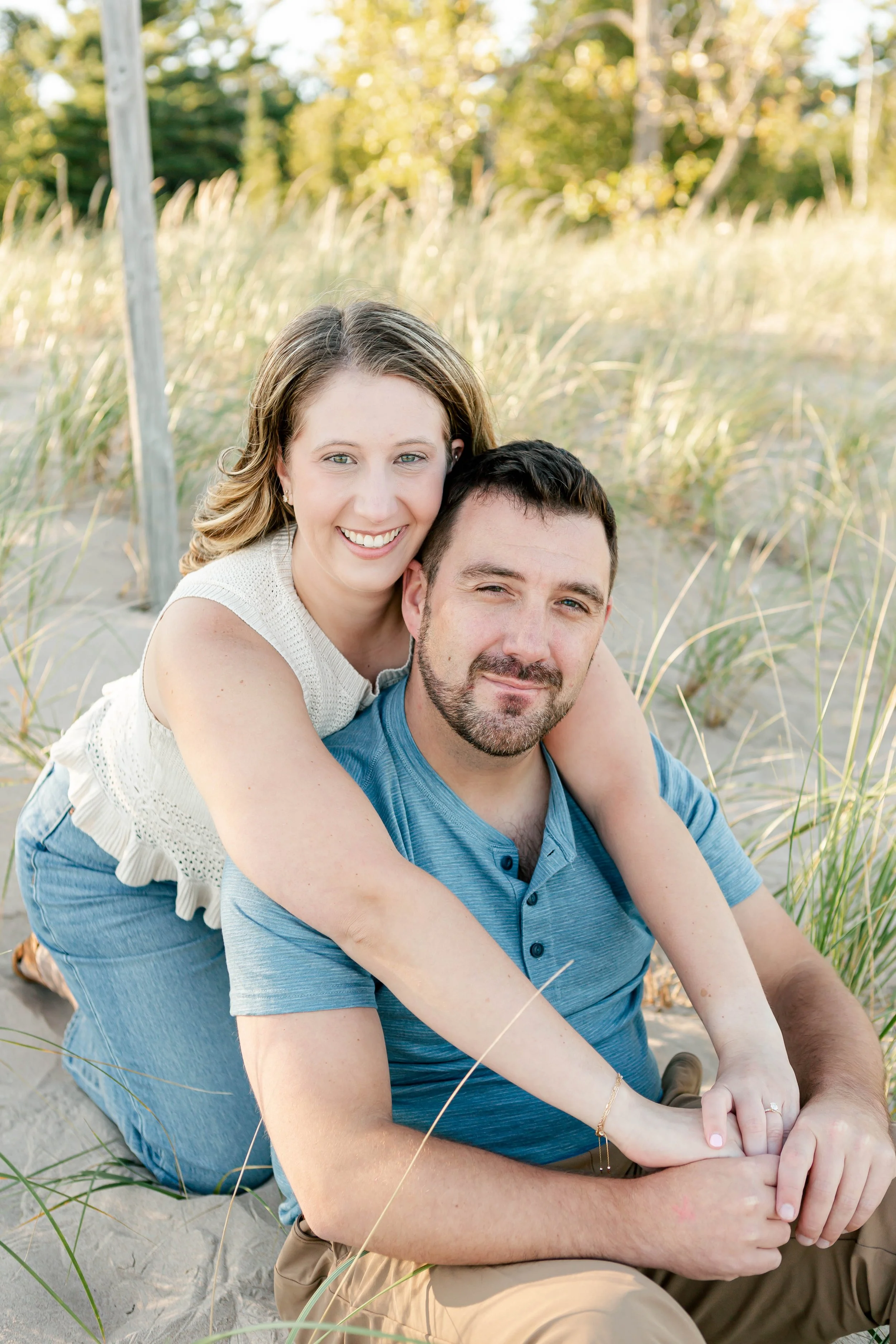 A smiling woman with light brown hair hugging a serious-looking man with dark hair and a beard at the beach, surrounded by tall grass and trees in the background.