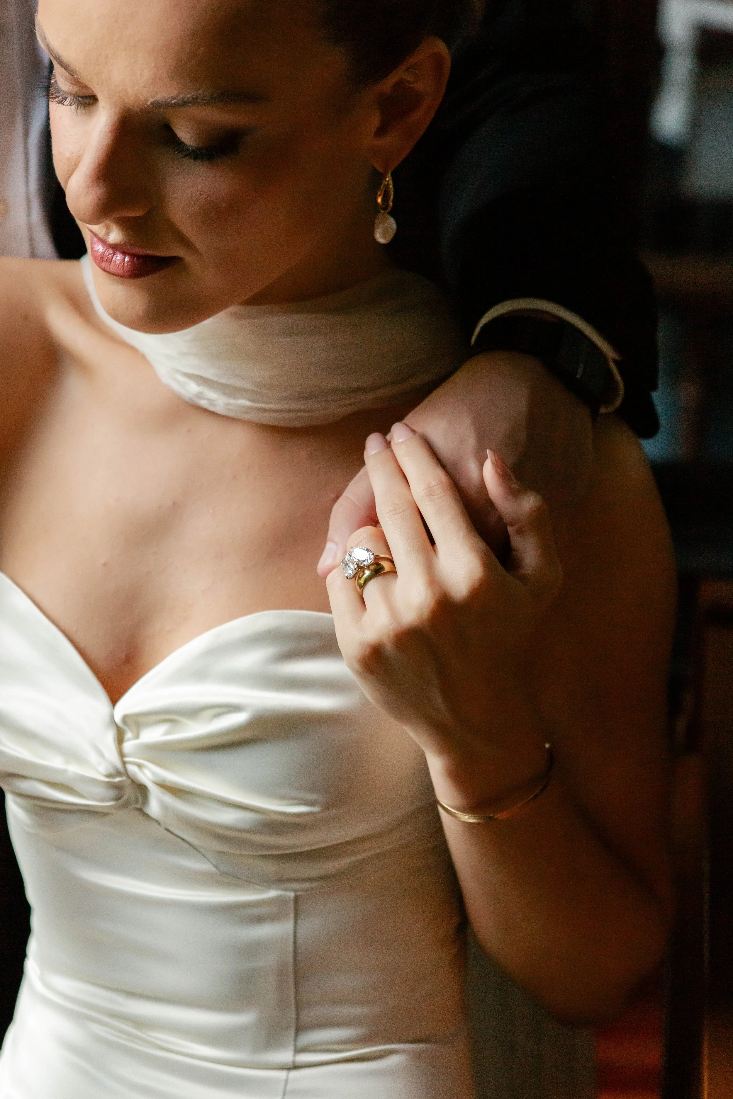 Close-up of a woman wearing a cream-colored satin dress, with a large ring on her finger, and a man holding her shoulder from behind.