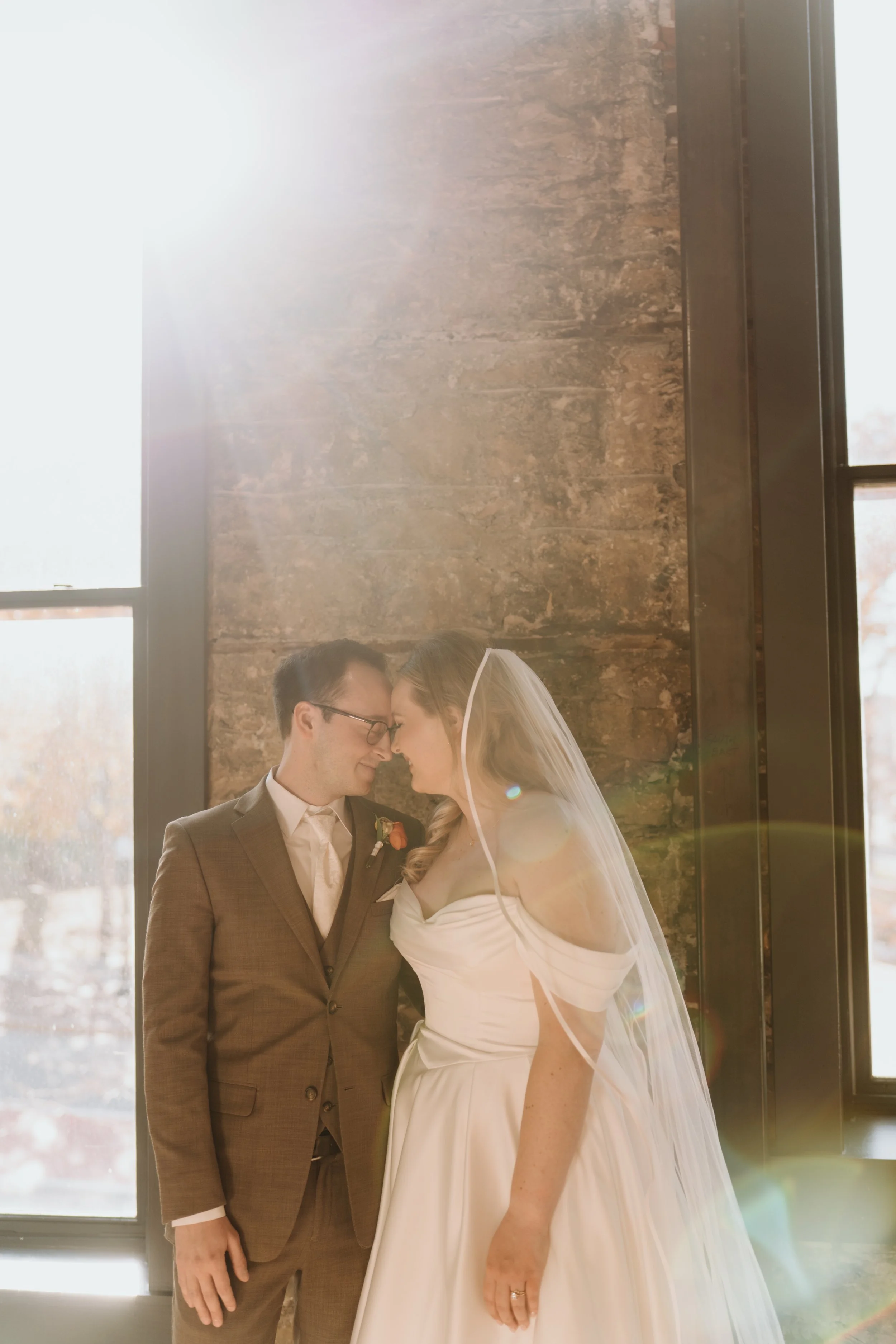 A bride and groom standing close together indoors, touching foreheads and smiling, with large window behind them allowing sunlight to stream in, revealing a brick wall and autumn trees outside.