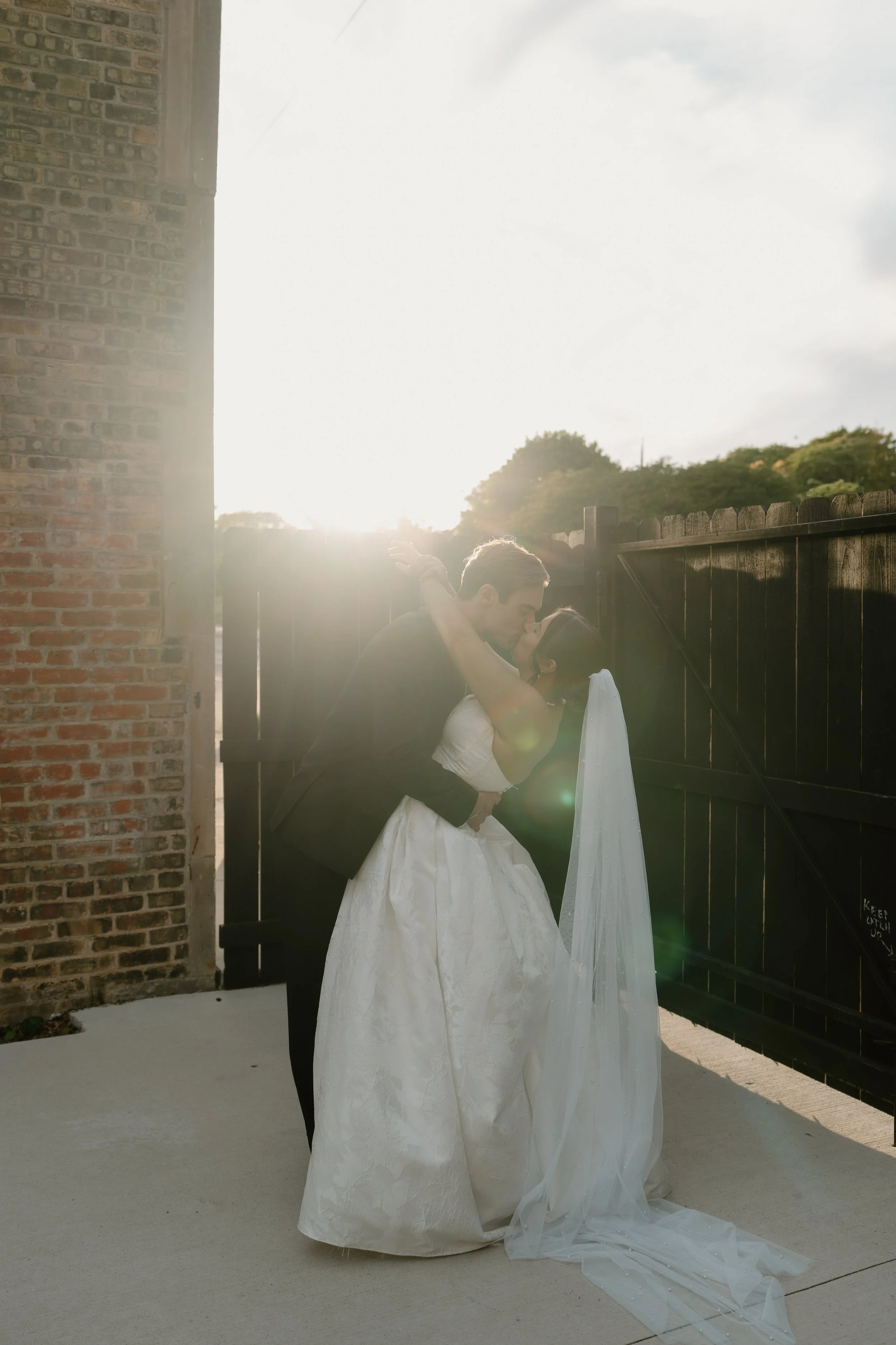 A couple kissing outdoors, dressed in wedding attire, with the groom in a black suit and the bride in a white wedding gown and veil, standing by a black wooden fence with trees in the background and bright sunlight.