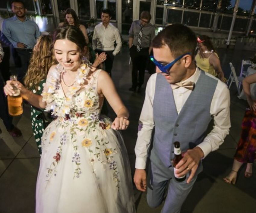 Young woman in a floral wedding dress dancing at a party, holding a drink, smiling. Young man in a gray vest, beige shirt, bow tie, and sunglasses dancing nearby, holding a beer. Several other people in semi-formal attire are in the background, enjoying the event in a bright, indoor venue with large windows.