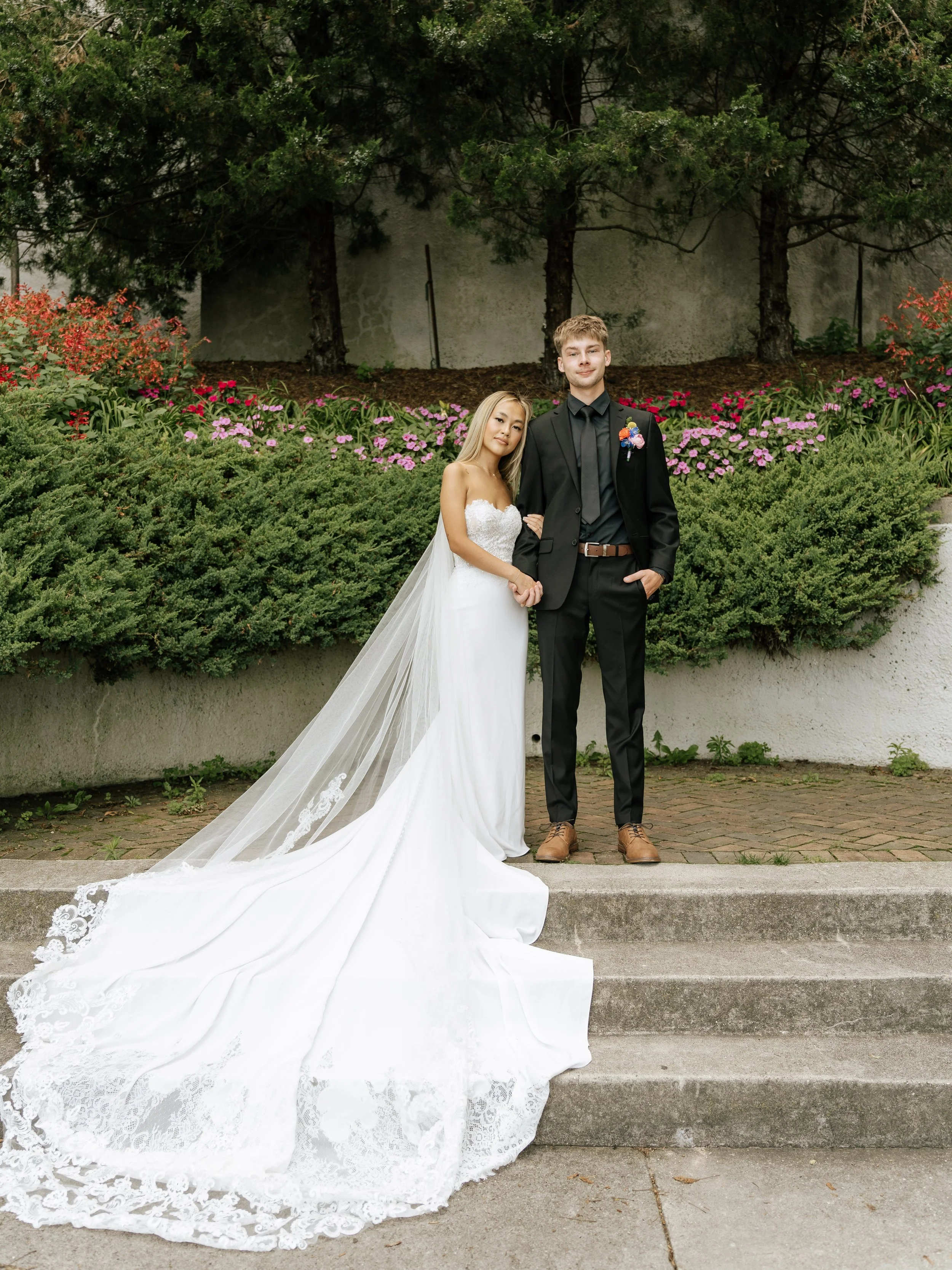 A bride and groom standing together outdoors in front of greenery and flowering bushes. The bride is wearing a white wedding gown with a long train and veil, and the groom is in a black suit and brown shoes.