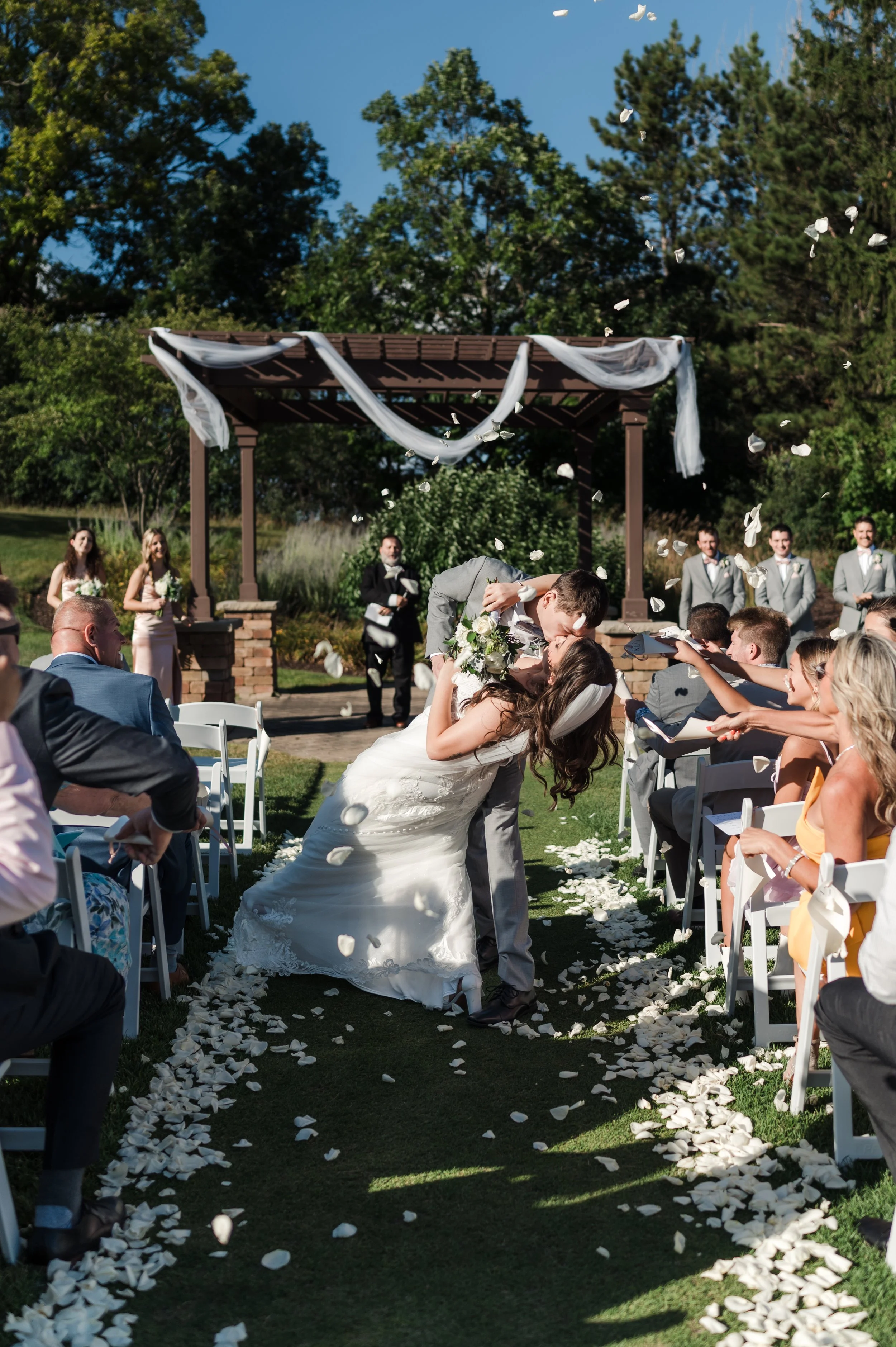 A newlywed couple shares a kiss during their outdoor wedding ceremony, surrounded by friends and family, with petals falling around them on a sunny day.