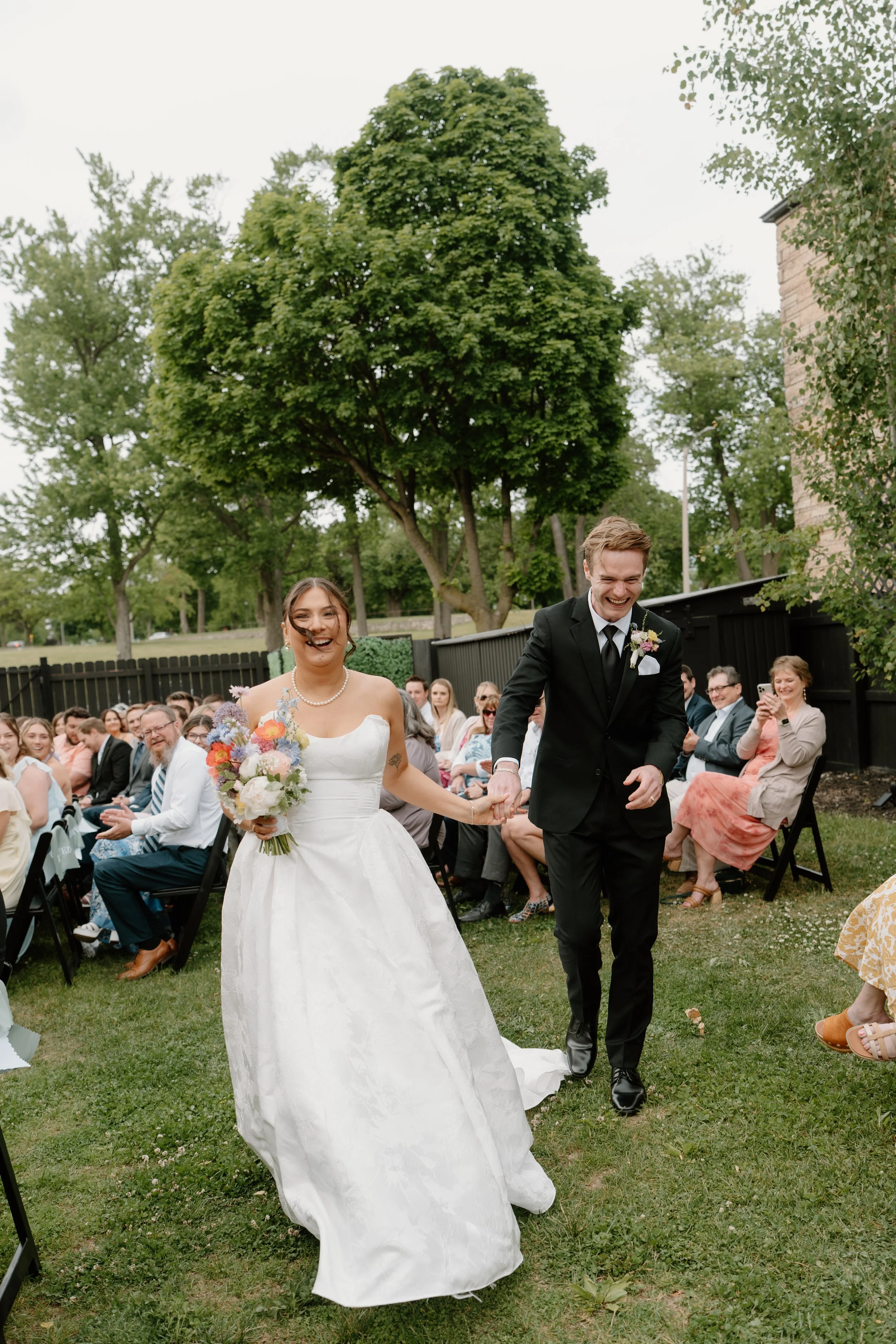 A bride and groom walking hand in hand outdoors during a wedding ceremony, with guests seated on chairs around them, in a garden setting with green trees and a wooden fence.