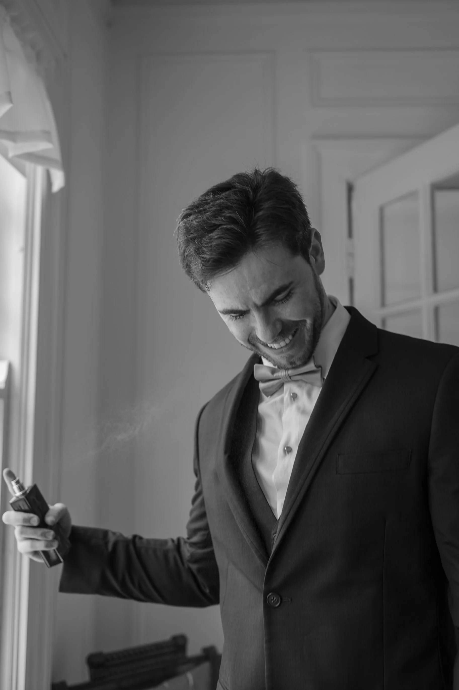 A man in a tuxedo, smiling and holding a tube of lipstick in front of a window.