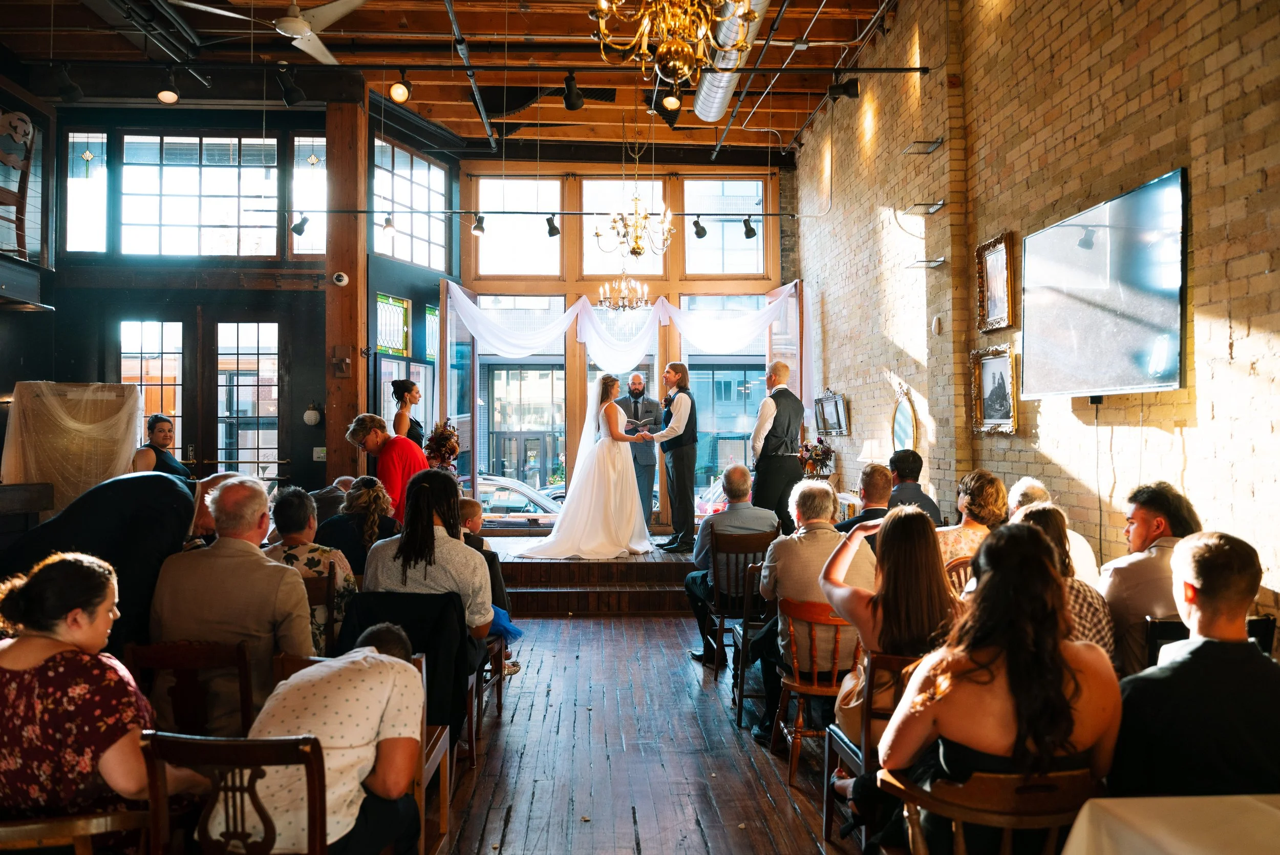A wedding ceremony taking place inside a spacious, brick-walled venue with large windows, chandeliers, and drapery. The bride and groom stand facing each other with officiant. Guests are seated, watching the ceremony. The room has an industrial style