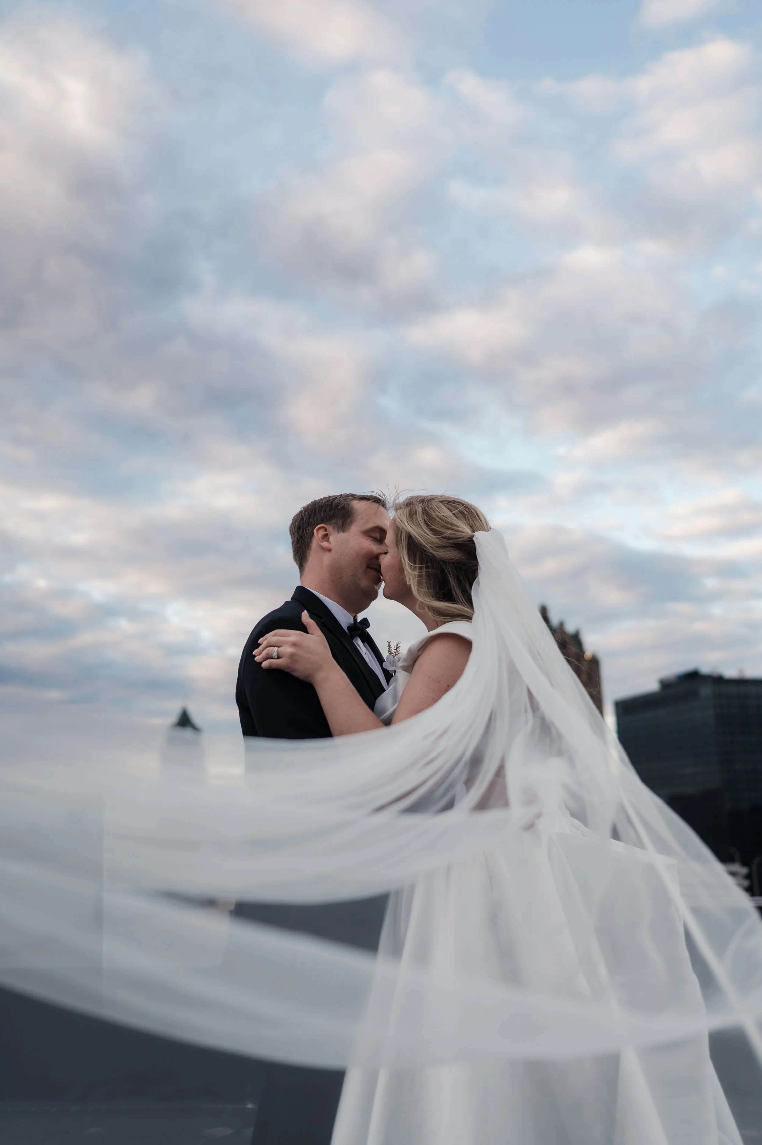 A newlywed couple sharing a kiss on a rooftop with a city skyline and cloudy sky in the background, the bride wearing a white veil and wedding gown, and the groom in a black tuxedo.