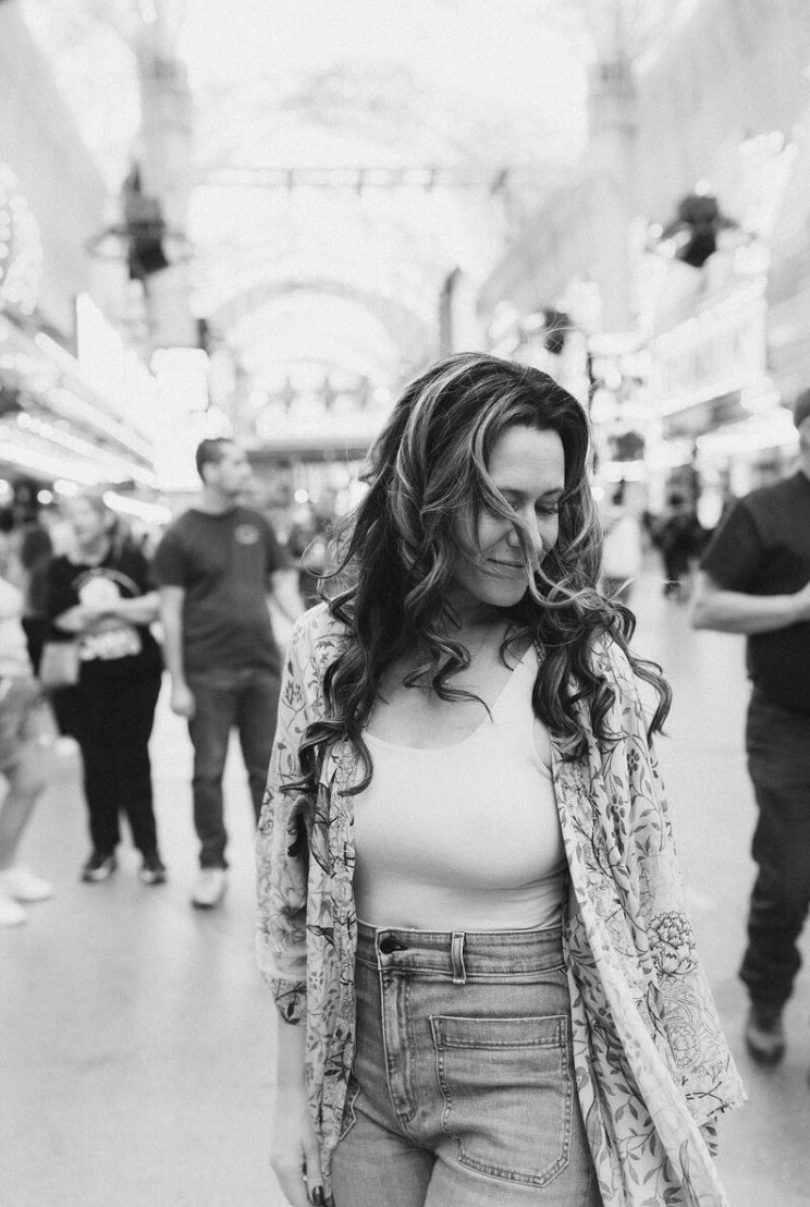 A woman with long, wavy hair standing in a busy indoor marketplace or arcade, looking down and smiling with her eyes closed, wearing a white top, faded high-waisted jeans, and a floral patterned cardigan.