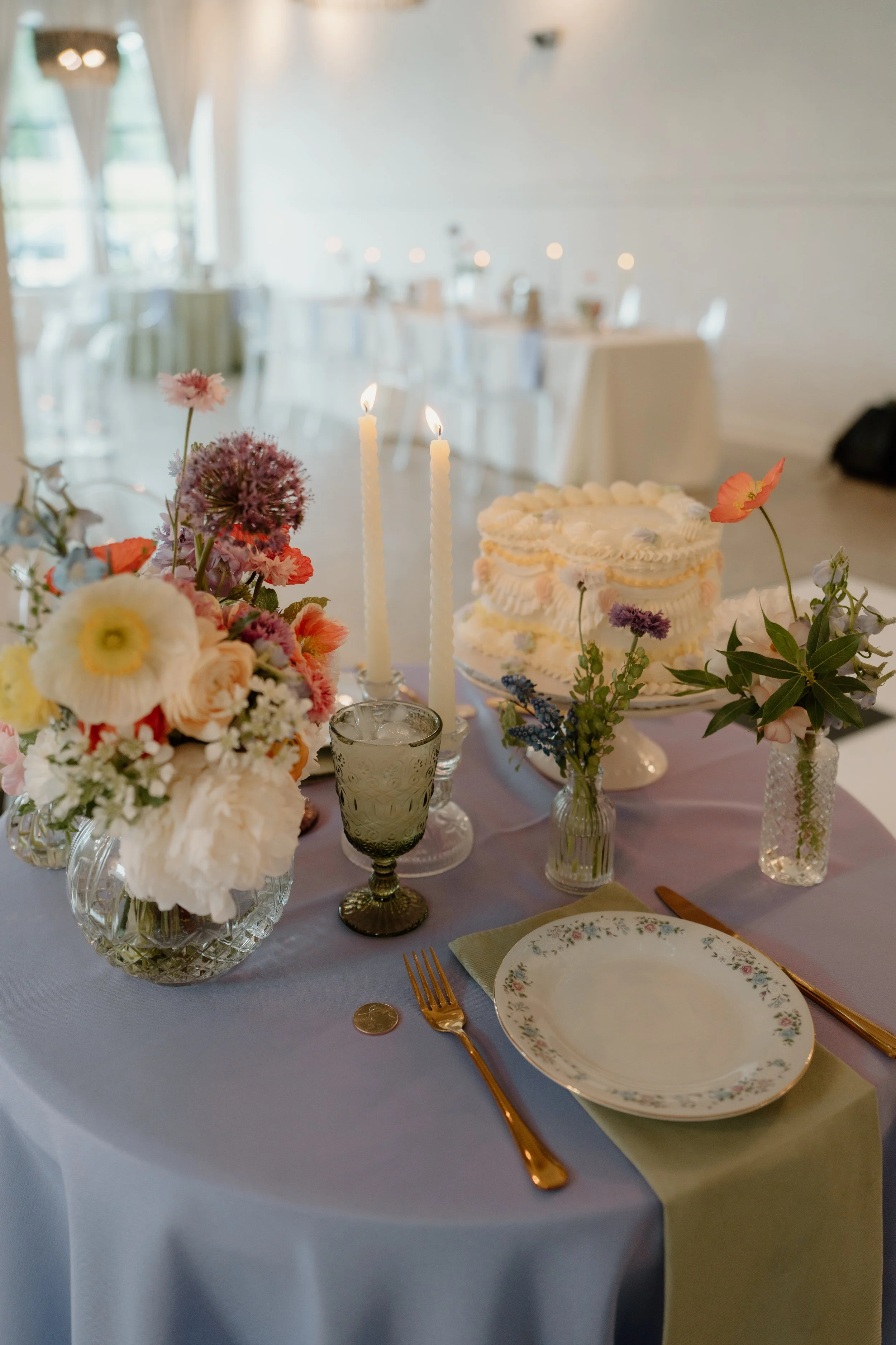 A decorated table at a celebration with floral arrangements, a white cake with pastel decorations, lit candles, and antique-style glassware and dishware, set in a well-lit room with large windows and draped curtains.