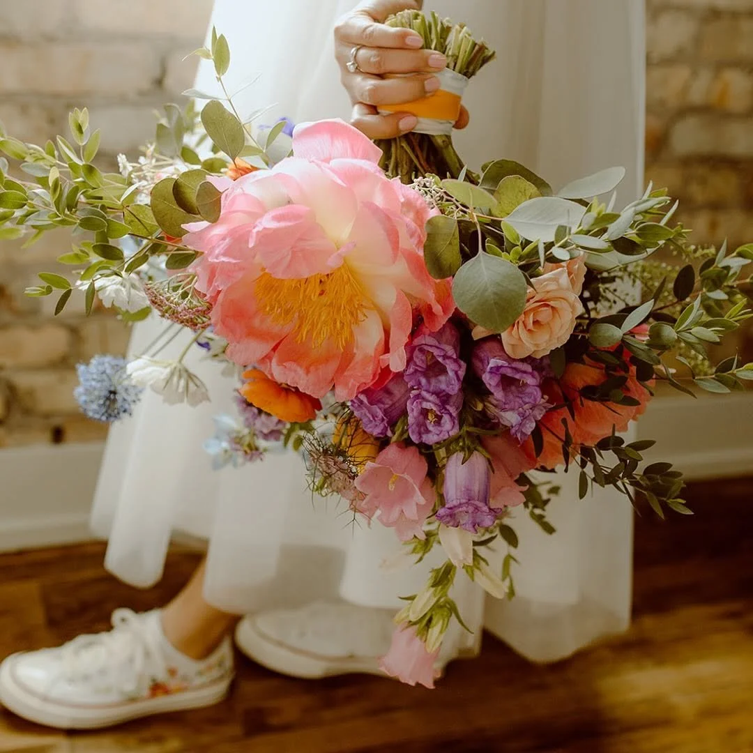 Person holding a colorful bouquet of flowers, with a white dress and sneakers, standing near a brick wall.