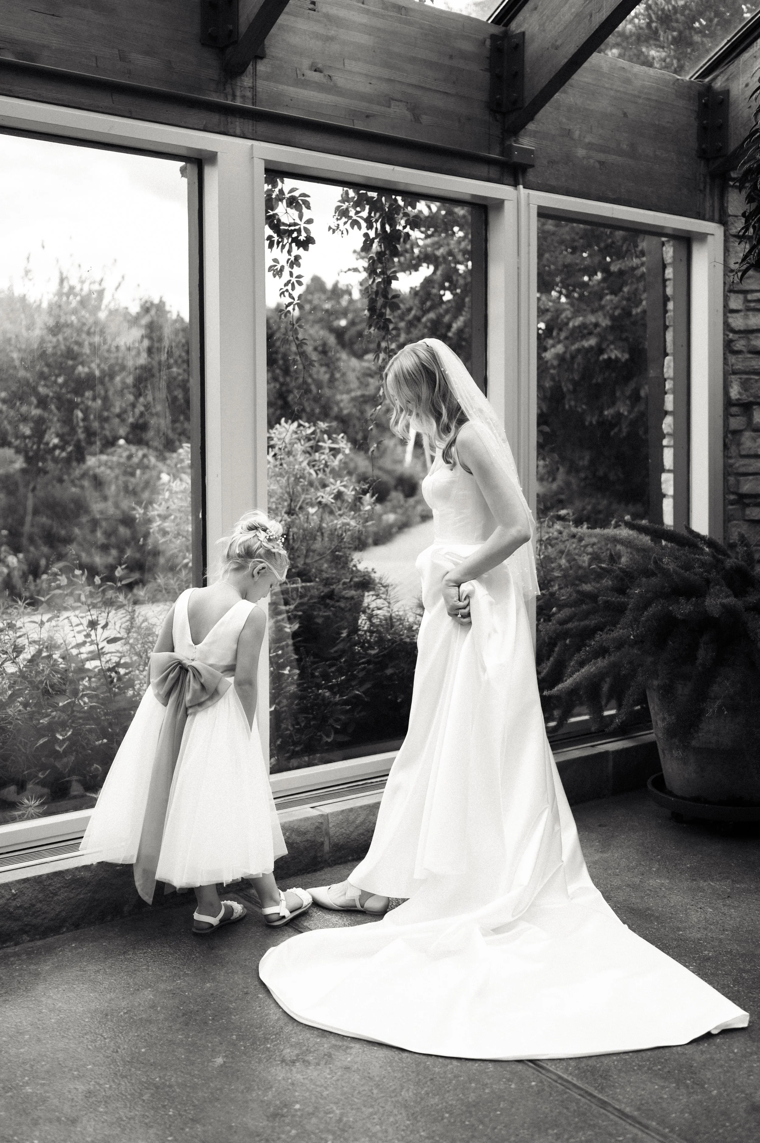 A bride in a white wedding dress and veil standing next to a young girl in a white dress with a large bow, both looking down at their shoes inside a building with large windows showing outdoor greenery.