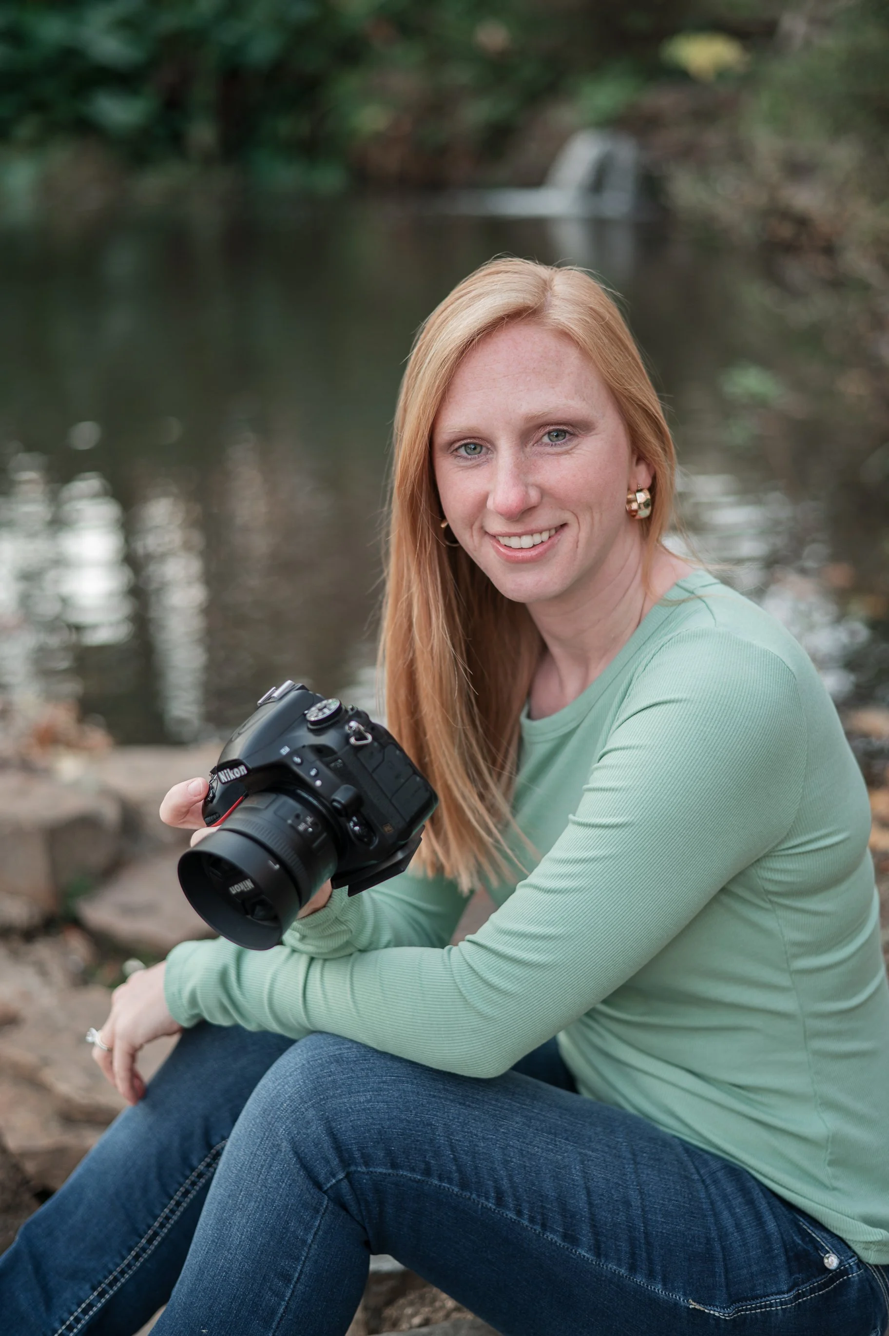 A woman with red hair and a green long-sleeve shirt sitting outdoors near a water feature, holding a camera, smiling at the camera.