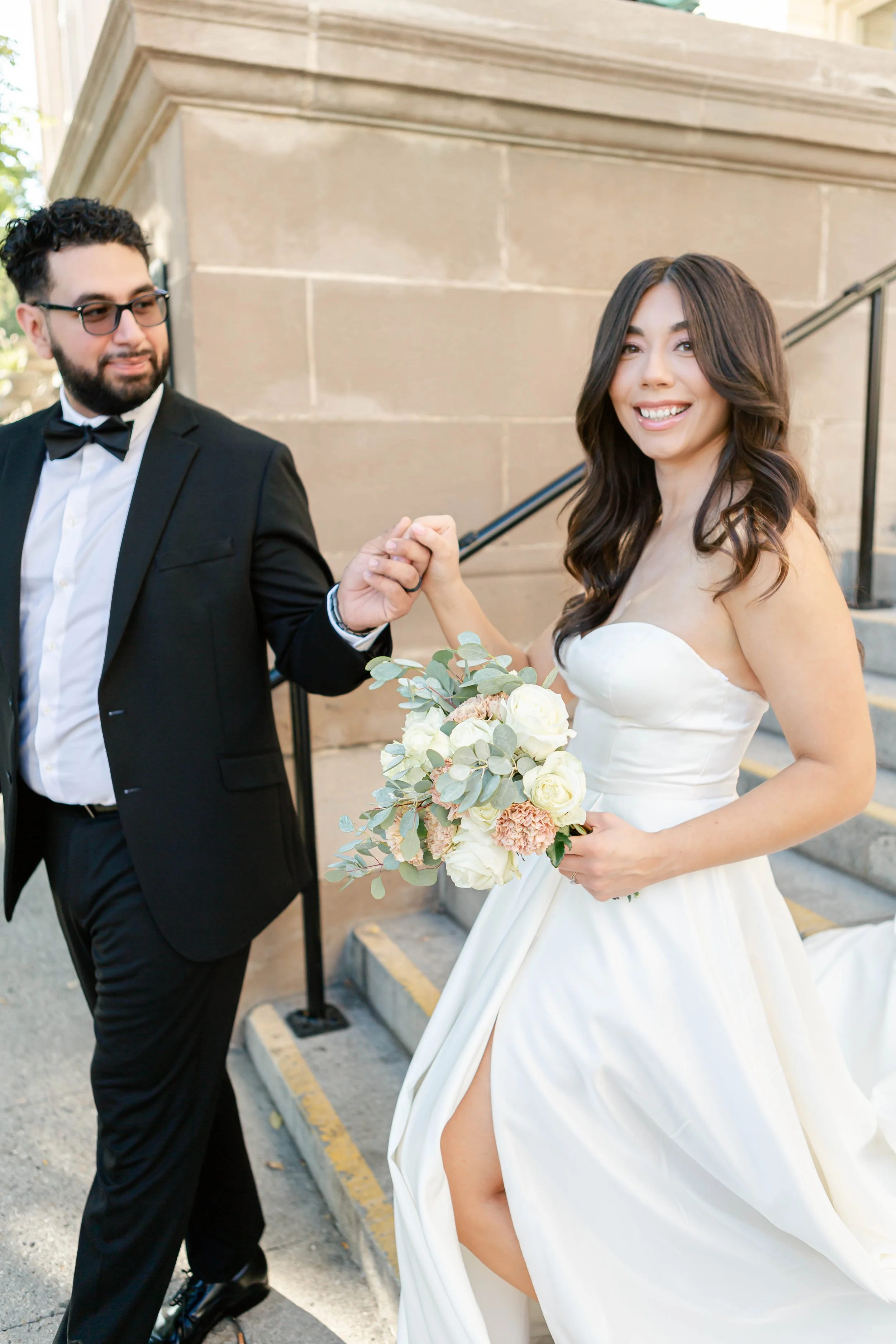 A newlywed couple, a man in a black tuxedo with glasses and a woman in a strapless white wedding gown, holding hands and smiling outside a building with stone steps. The woman holds a bouquet of white and pink flowers and greenery.