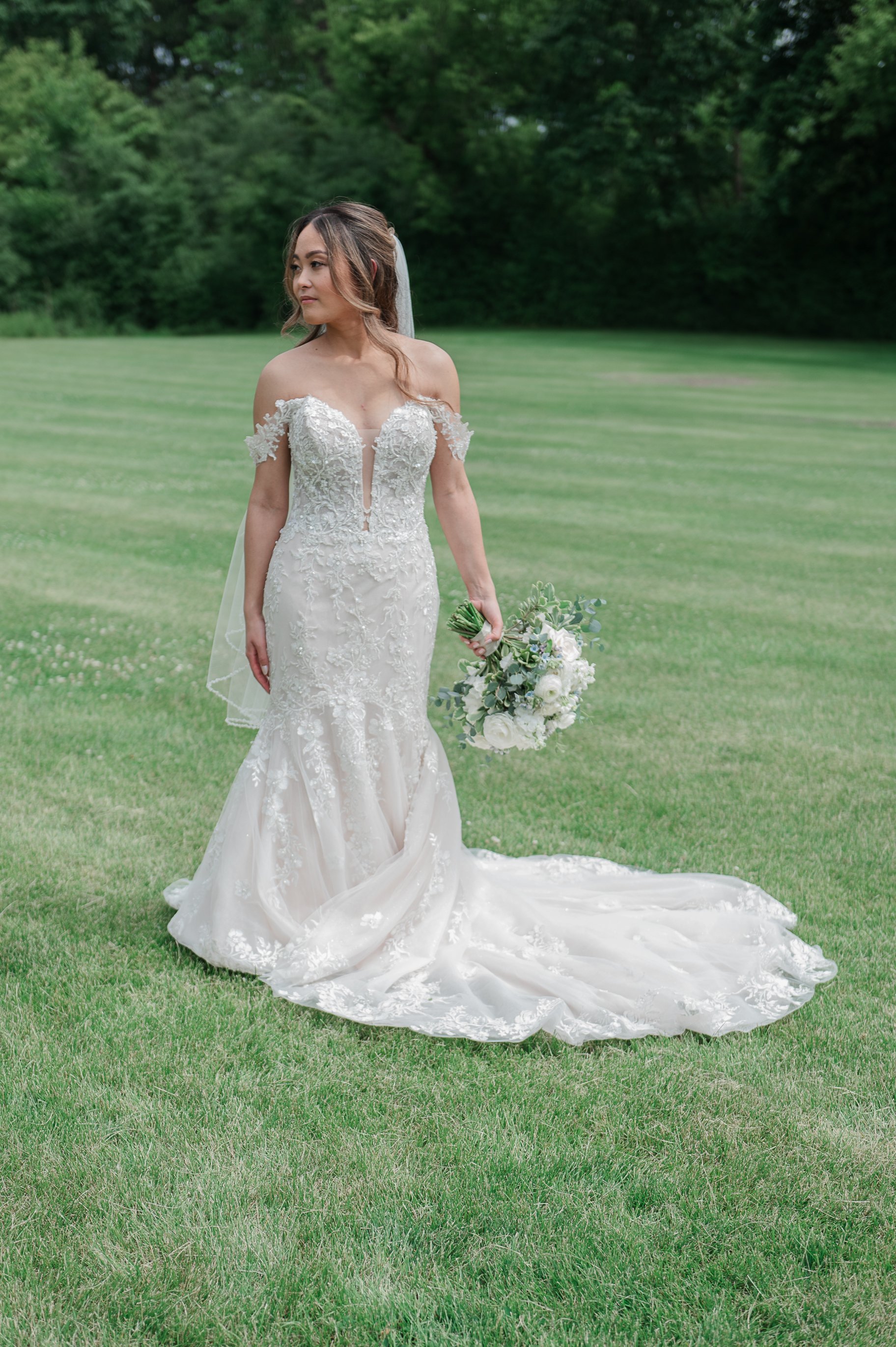 A bride in a white wedding gown holding a bouquet of white and purple flowers stands on a grassy field.
