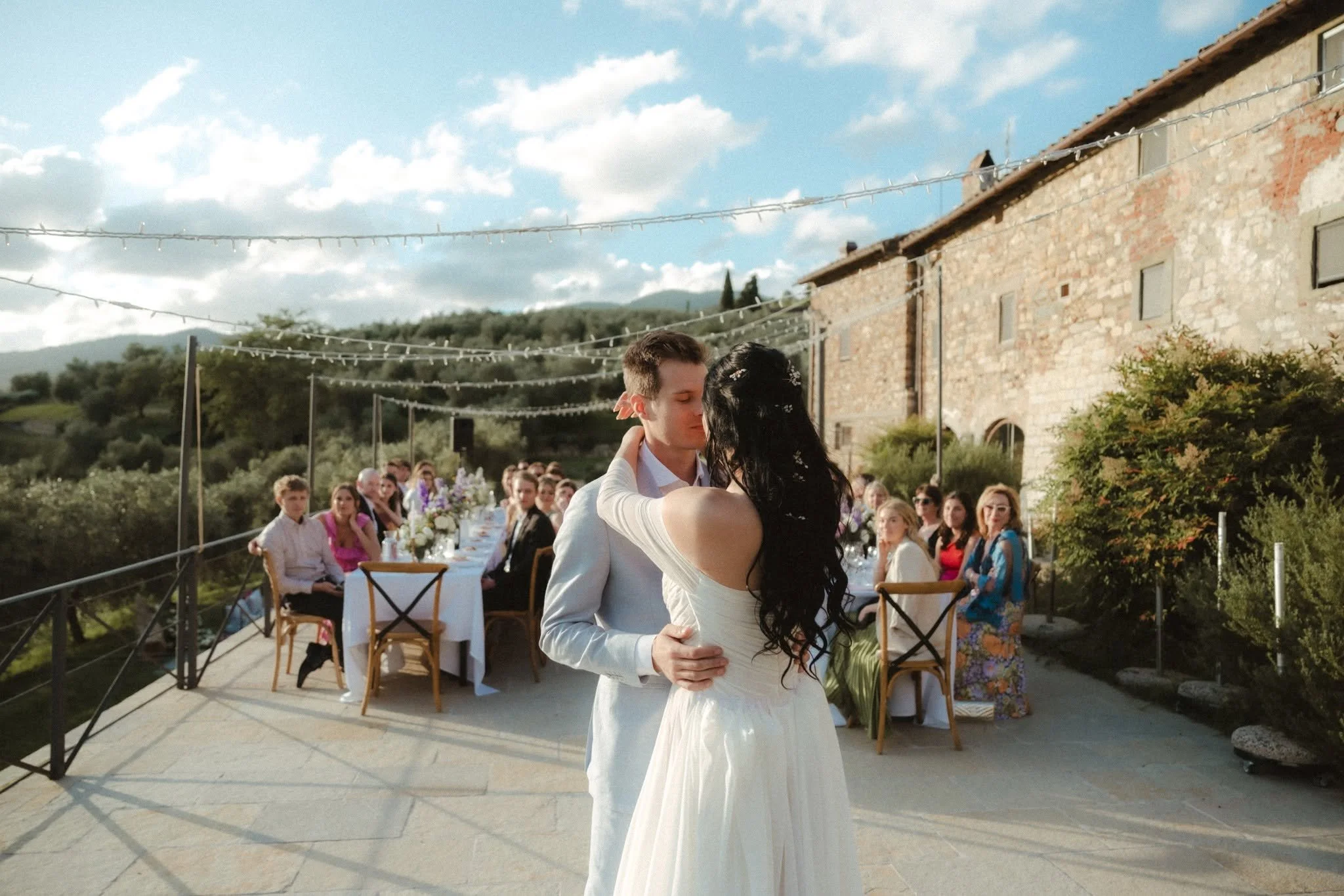 A bride and groom sharing a dance at an outdoor wedding reception during daytime. Guests are seated at a long table in the background, and the setting includes a rustic brick building and string lights overhead.