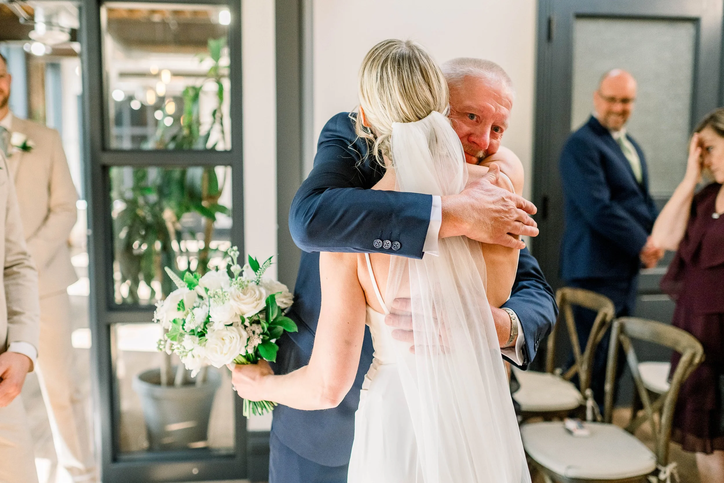 A wedding reception where a bride and groom are sharing a hug. The groom is holding a bouquet of white and green flowers. Other guests are present in the background, dressed in formal attire.