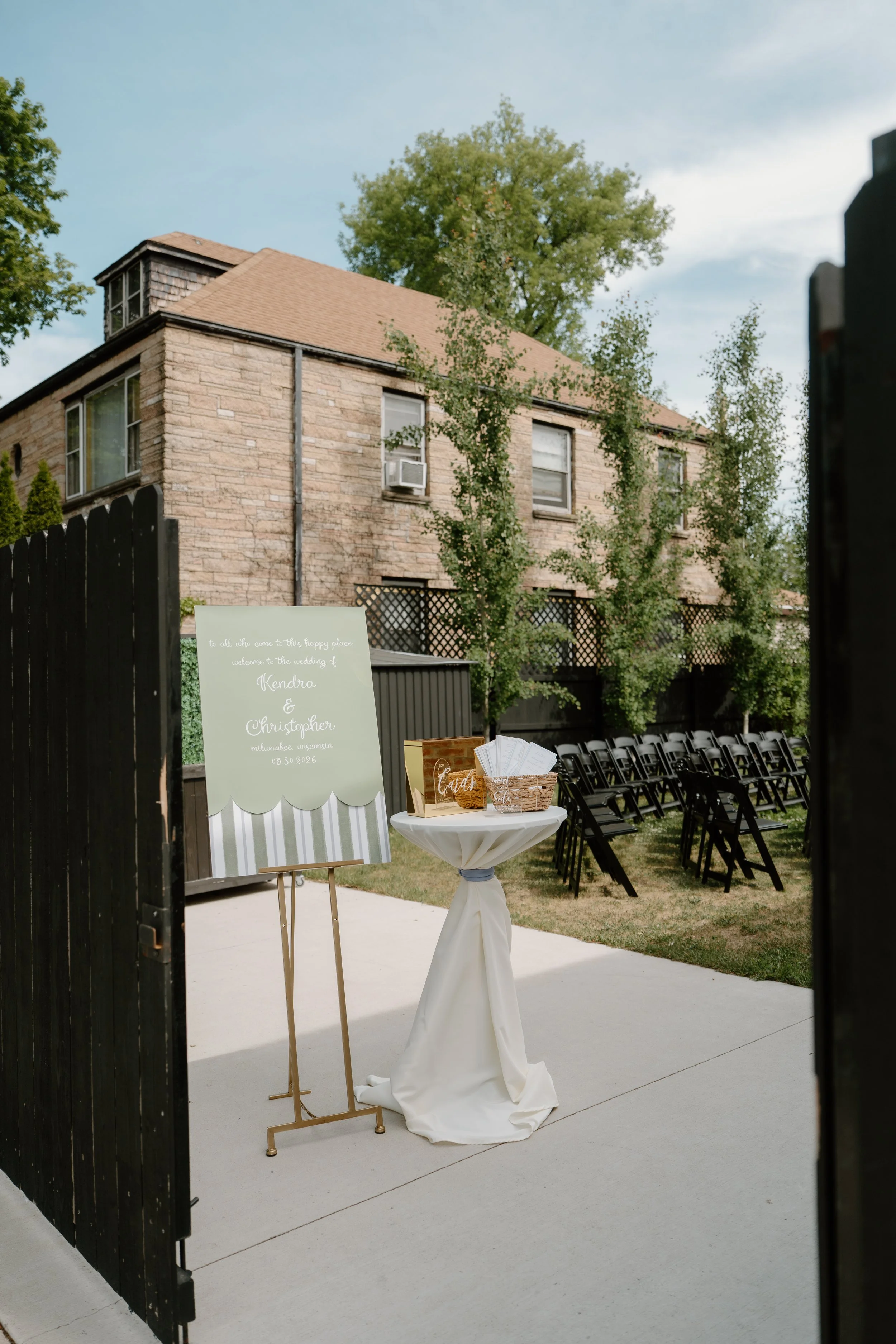 Outdoor wedding ceremony setup with black chairs, a sign welcoming guests named Kendra and Christopher, and a table with wedding programs and decorations, surrounded by trees and a brick building in the background.