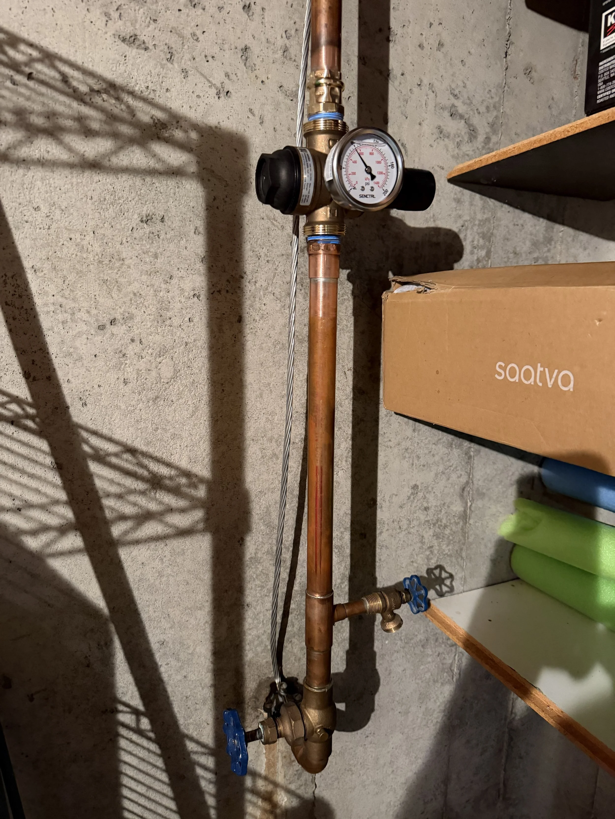 Copper plumbing pipes with a pressure gauge, shut-off valves, and fittings on a concrete wall in a basement or utility room.