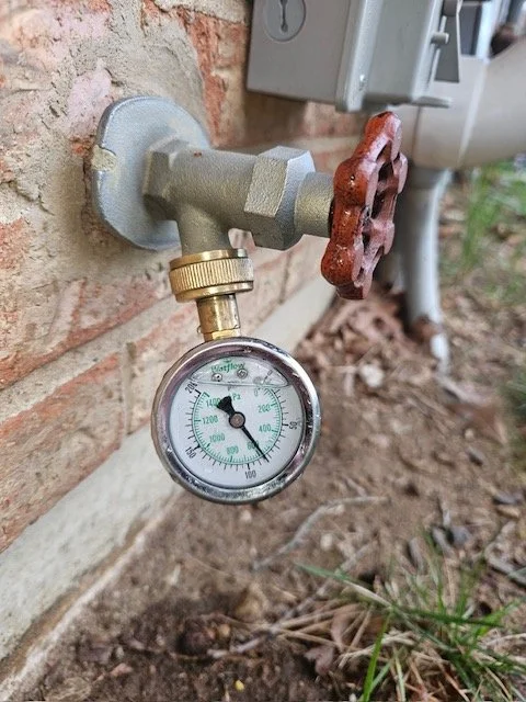 A close-up of a water supply valve with a pressure gauge attached, mounted on an exterior brick wall outside a building.