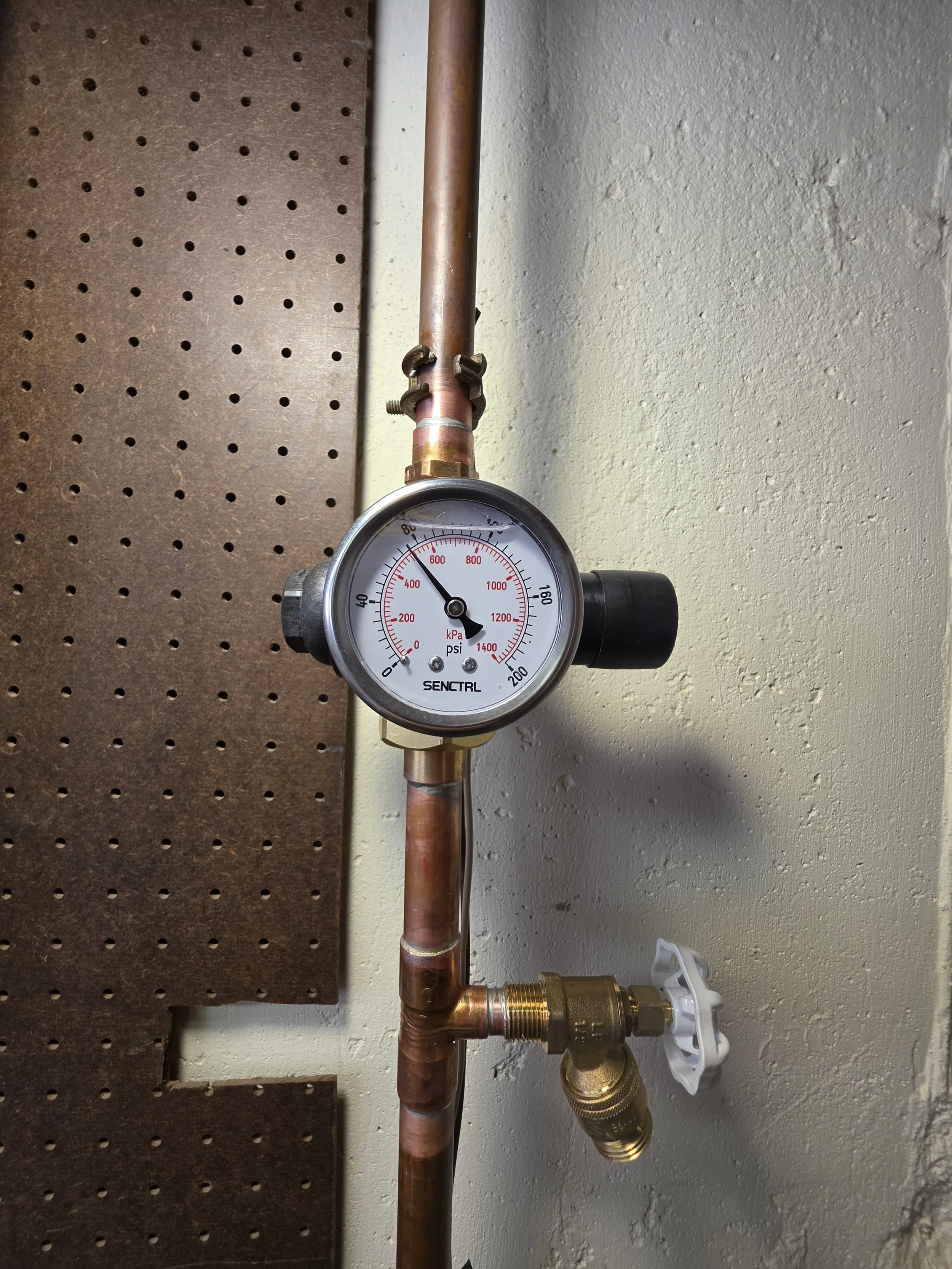 A pressure gauge connected to copper pipes and a valve on a wall.