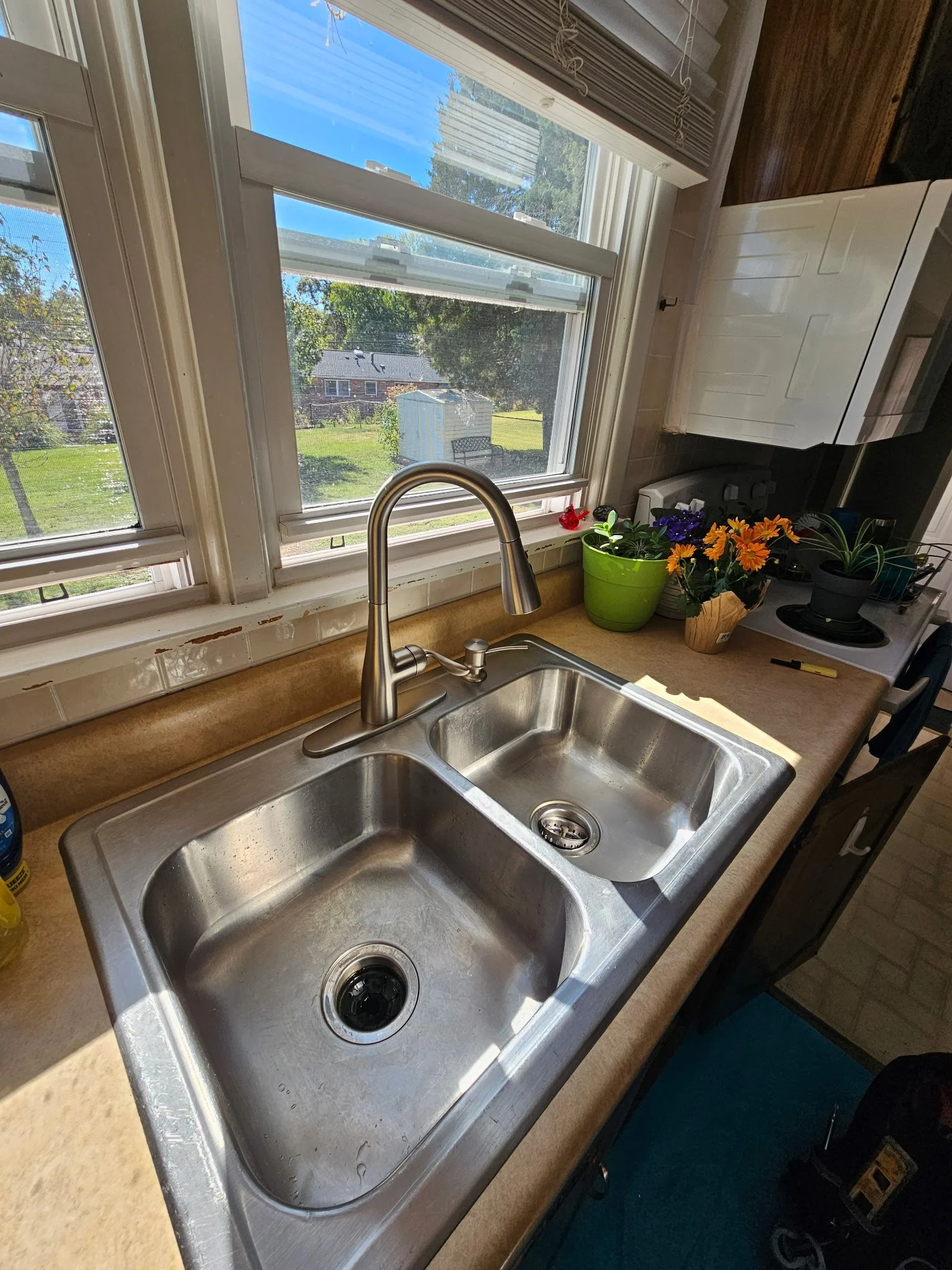View of a kitchen sink with two basins, a faucet, and a window showing a yard with trees, a bench, and a shed outside. On the countertop are potted plants and a cutting board.