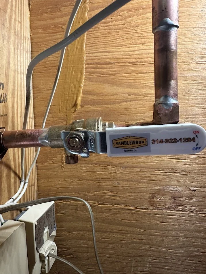 Pipes and a shut-off valve in a wooden wall, with a white handle labeled 'Ramblewood Plumbing Co.' and the contact number. The handle is in the 'Off' position.
