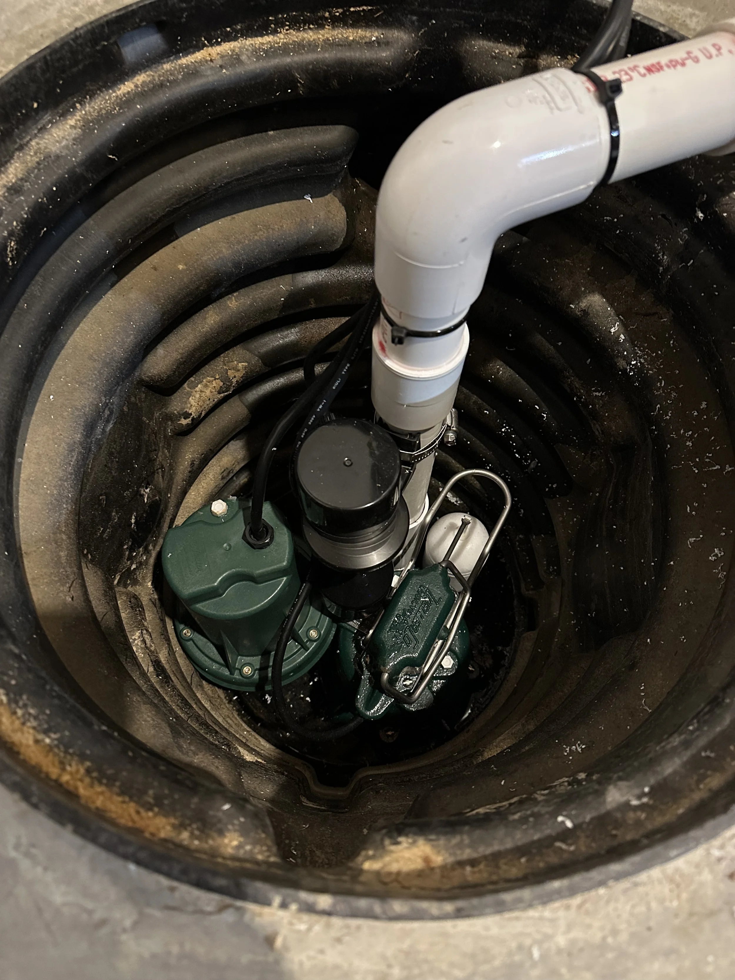 View inside a sump pump basin showing a submersible sump pump with thick electrical cord, white PVC discharge pipe, and surrounding basin walls.