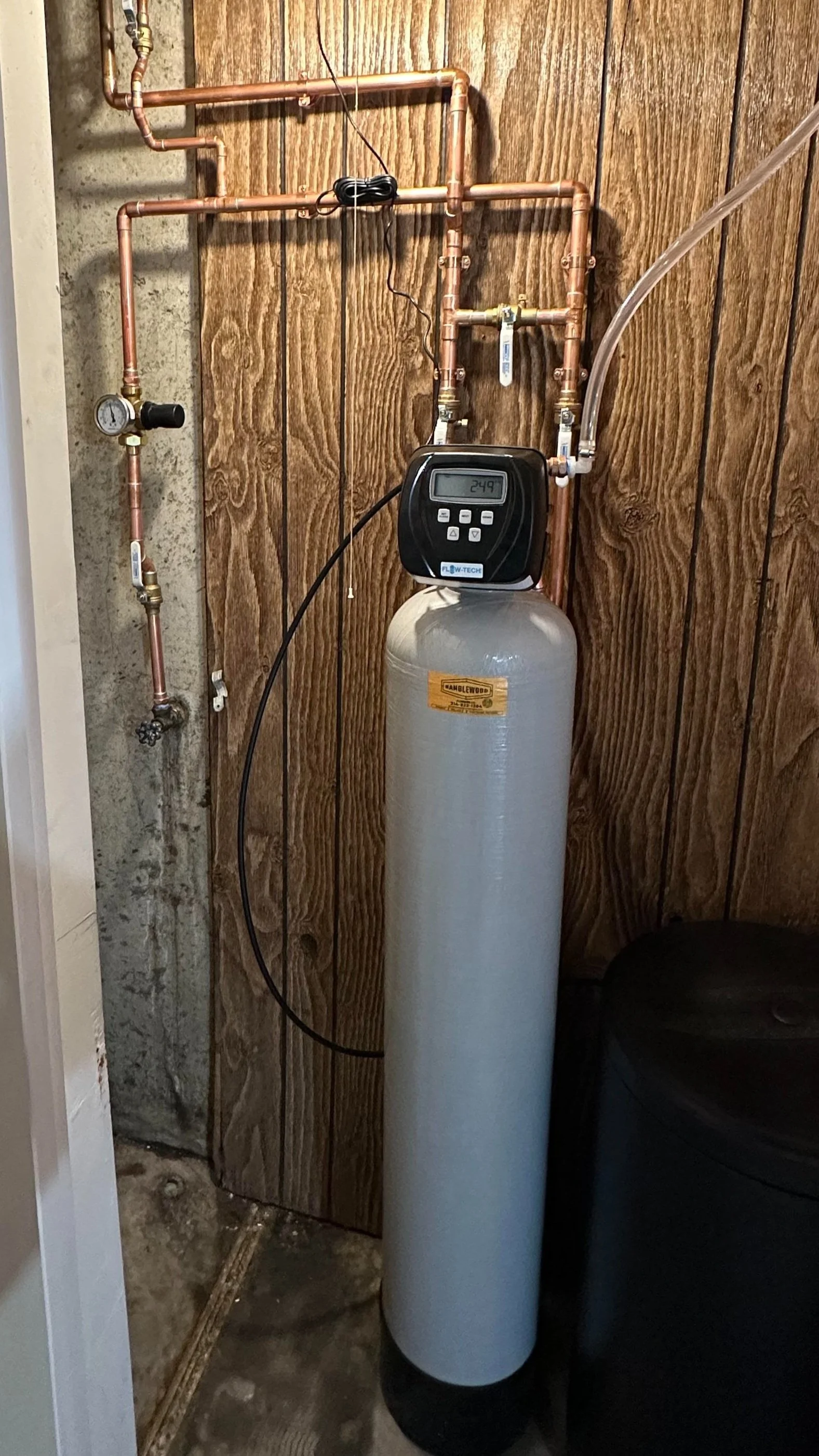 A water softener system with a control head displaying 24.9, copper piping, a pressure gauge, and tubing, installed against a wooden wall in a utility area.
