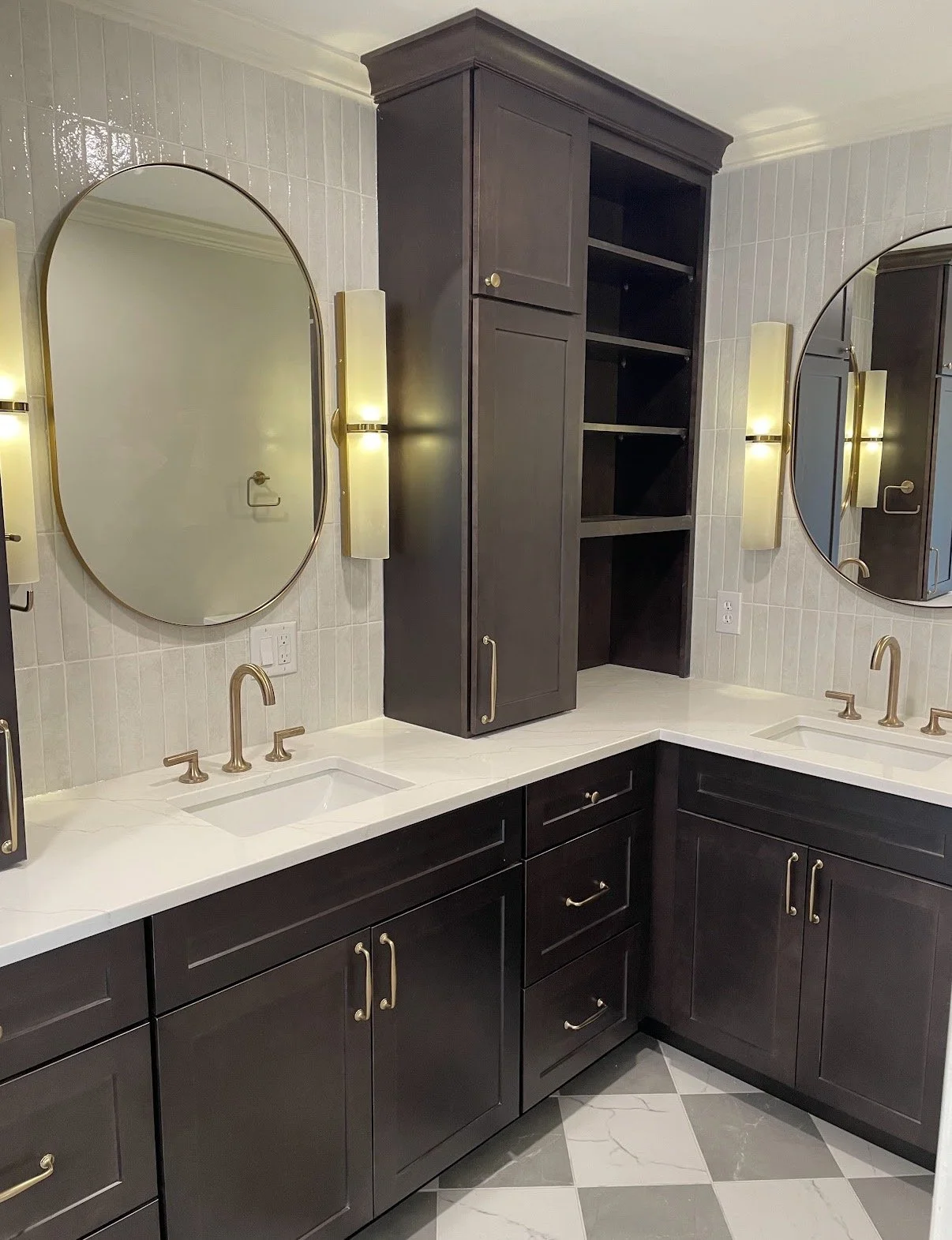 Bathroom with double vanity, dark wood cabinets, white countertop, oval mirrors, wall-mounted vertical light fixtures, and a tall dark wood cabinet in between the mirrors.