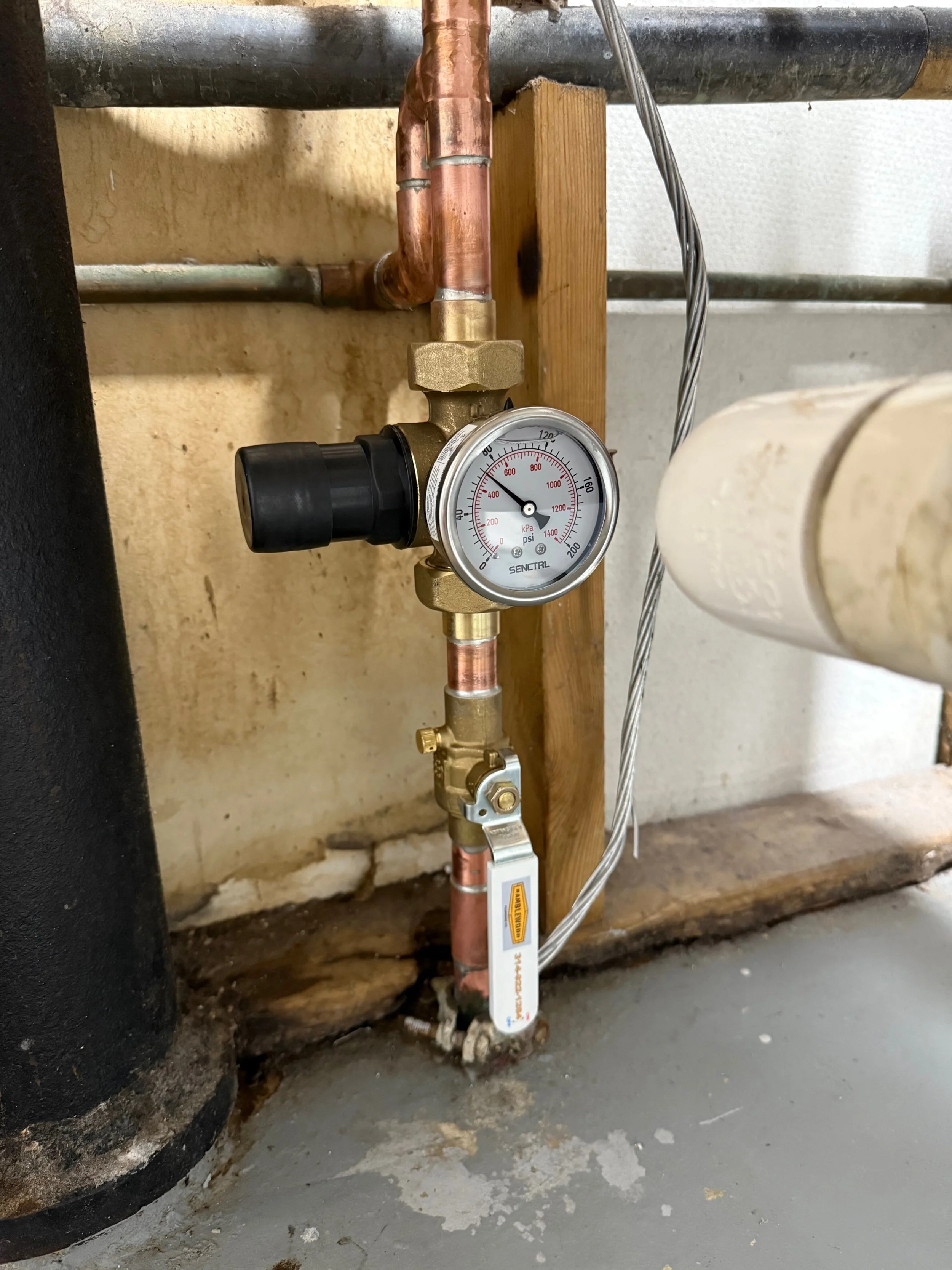 Close-up of a pressure gauge, shut-off valve, and copper piping in a plumbing system, mounted on a wall.