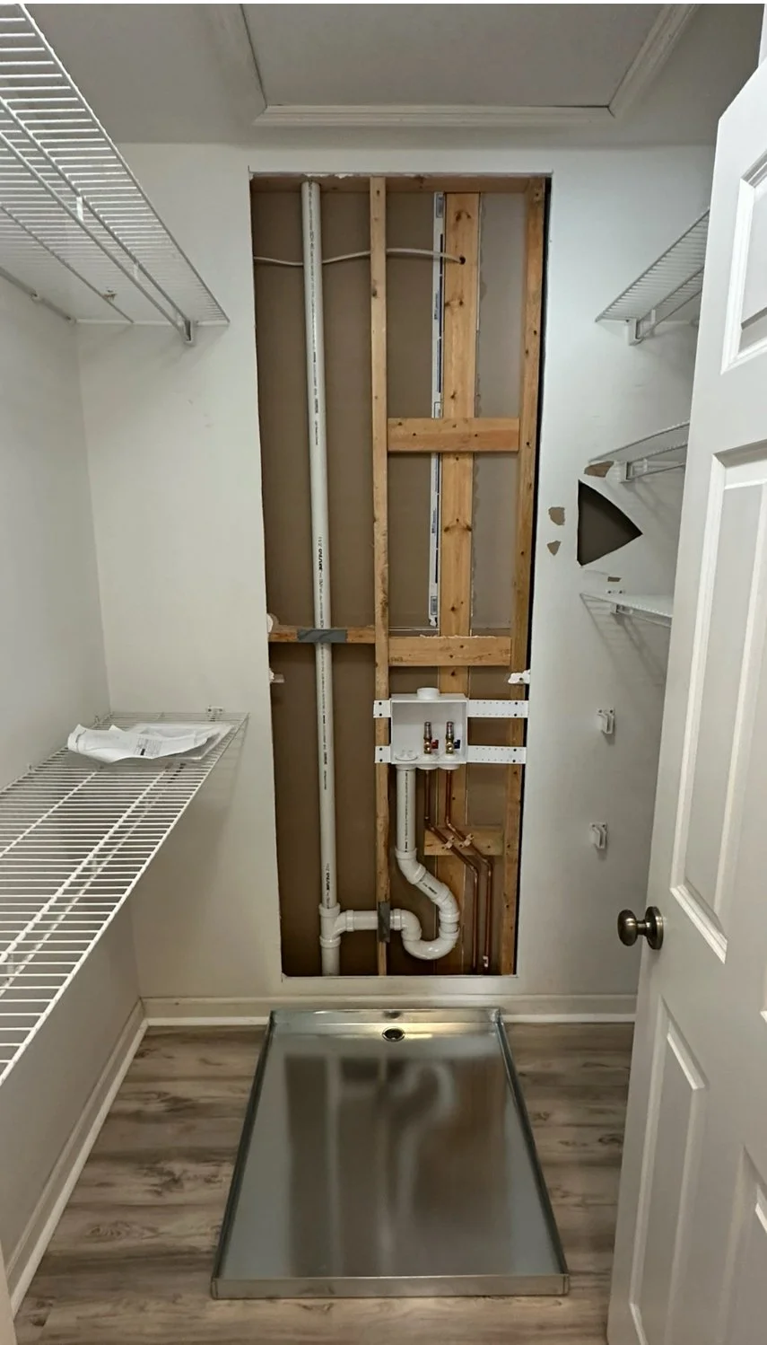 Empty laundry room with exposed wooden framing, plumbing pipes, wire shelving, and a stainless steel laundry pan on the floor.