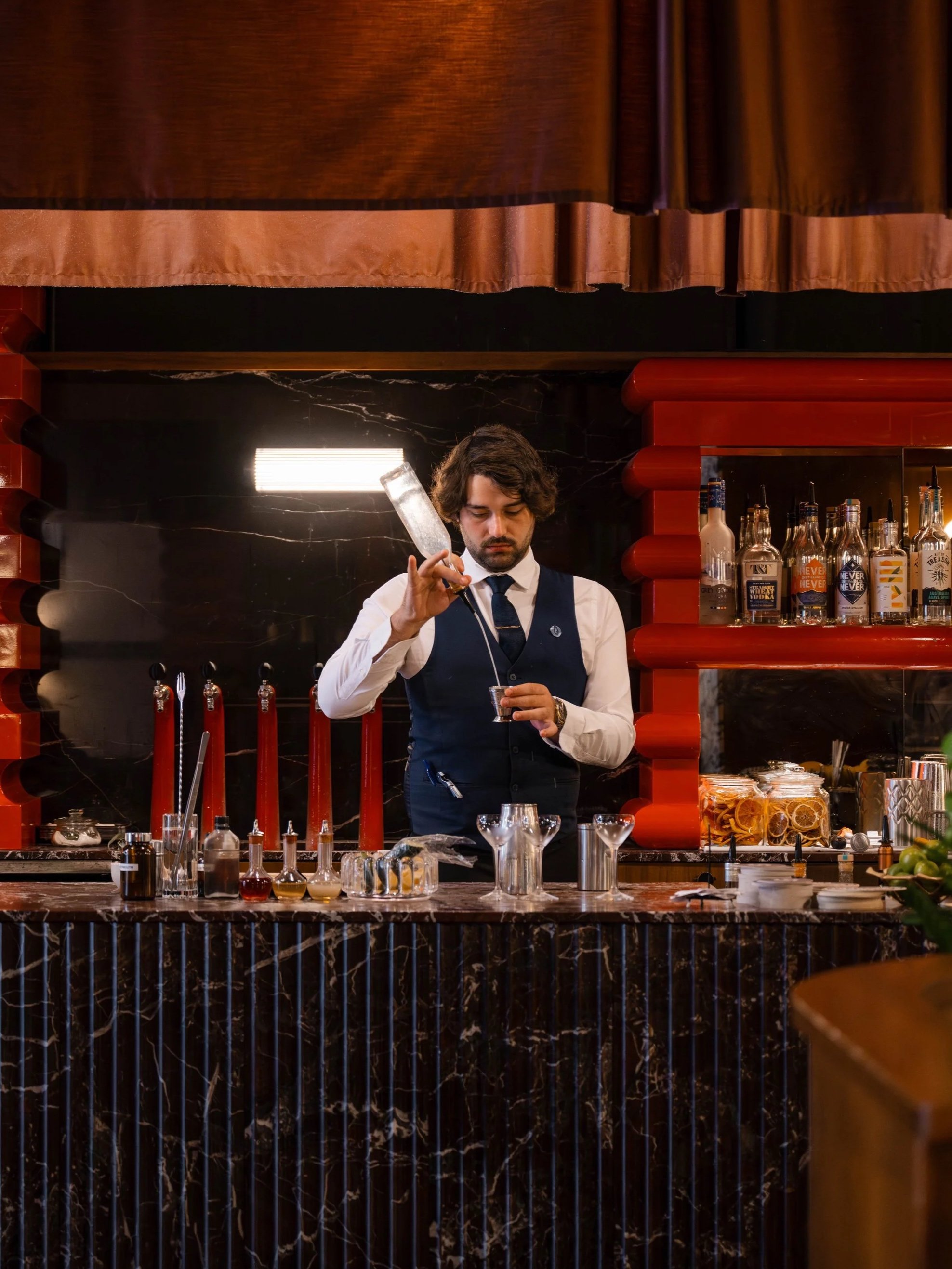 A bartender in formal attire prepares drinks at a bar with dark marble counter and red accents.