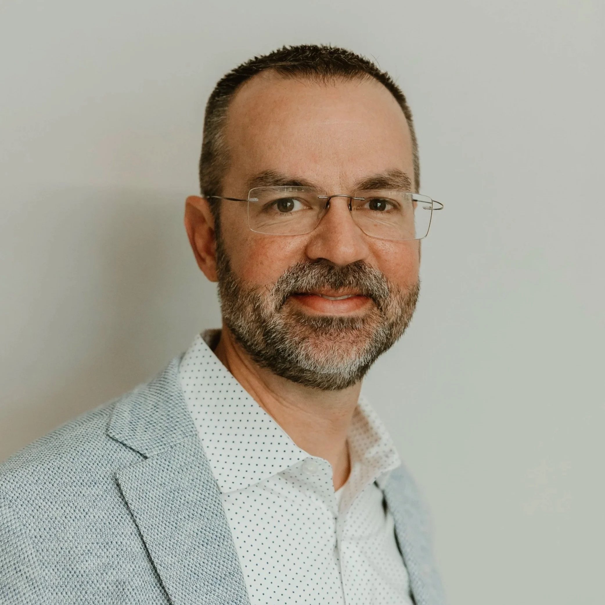 Close-up of a man with glasses, short brown hair, and a gray beard, wearing a white shirt with small black polka dots, standing against a plain light-colored wall.