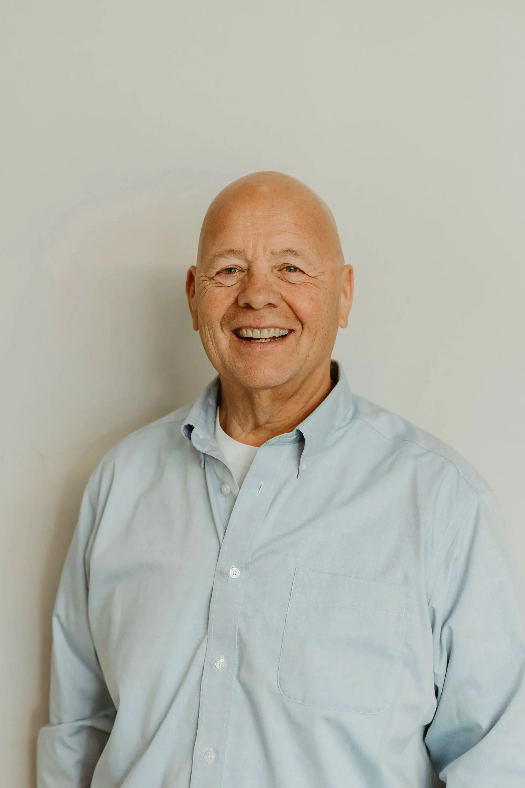 A smiling middle-aged man wearing a light blue collared shirt, standing against a plain white wall.