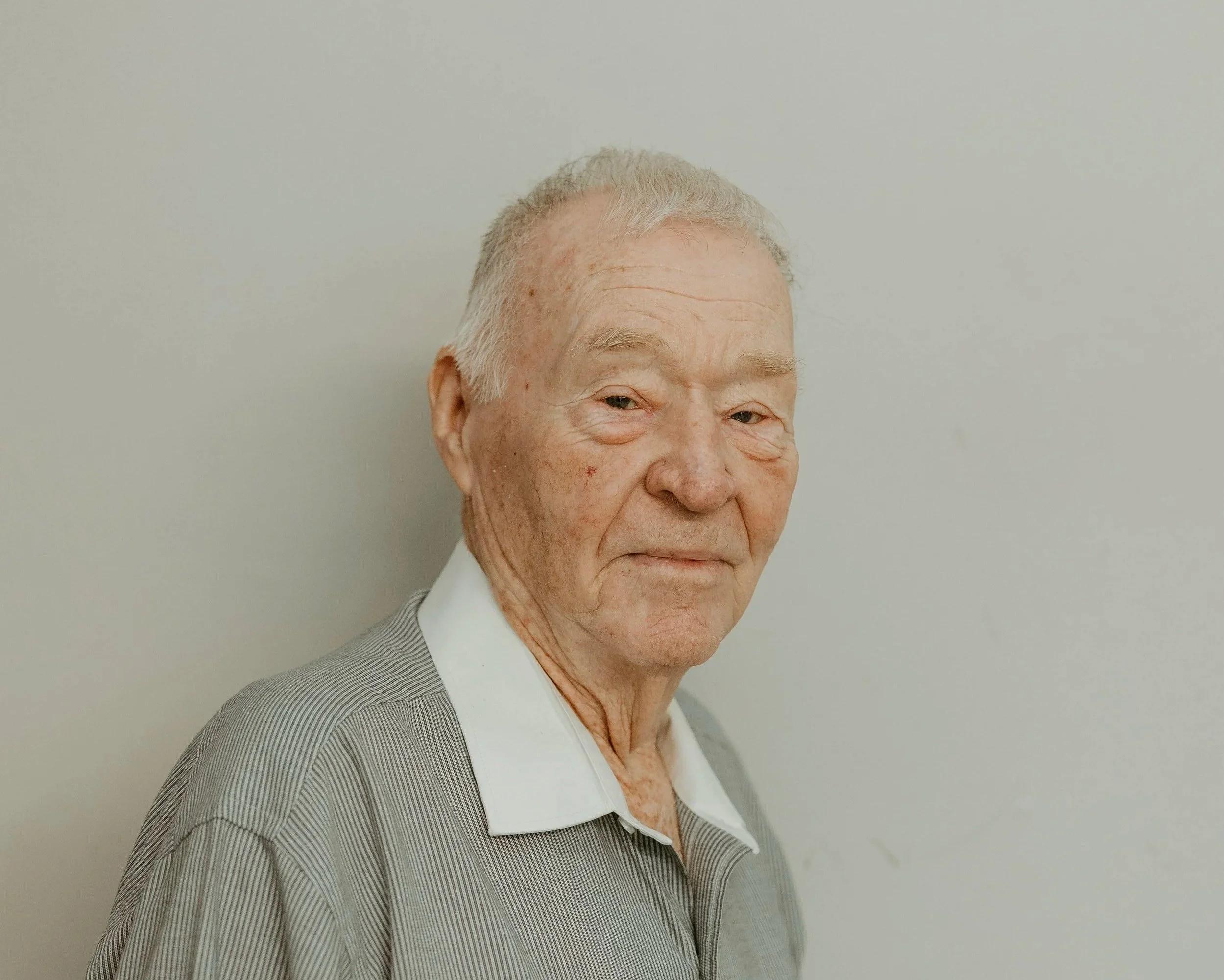 Close-up of an elderly man with light skin, gray hair, wearing a striped suit and white shirt against a plain light-colored background.
