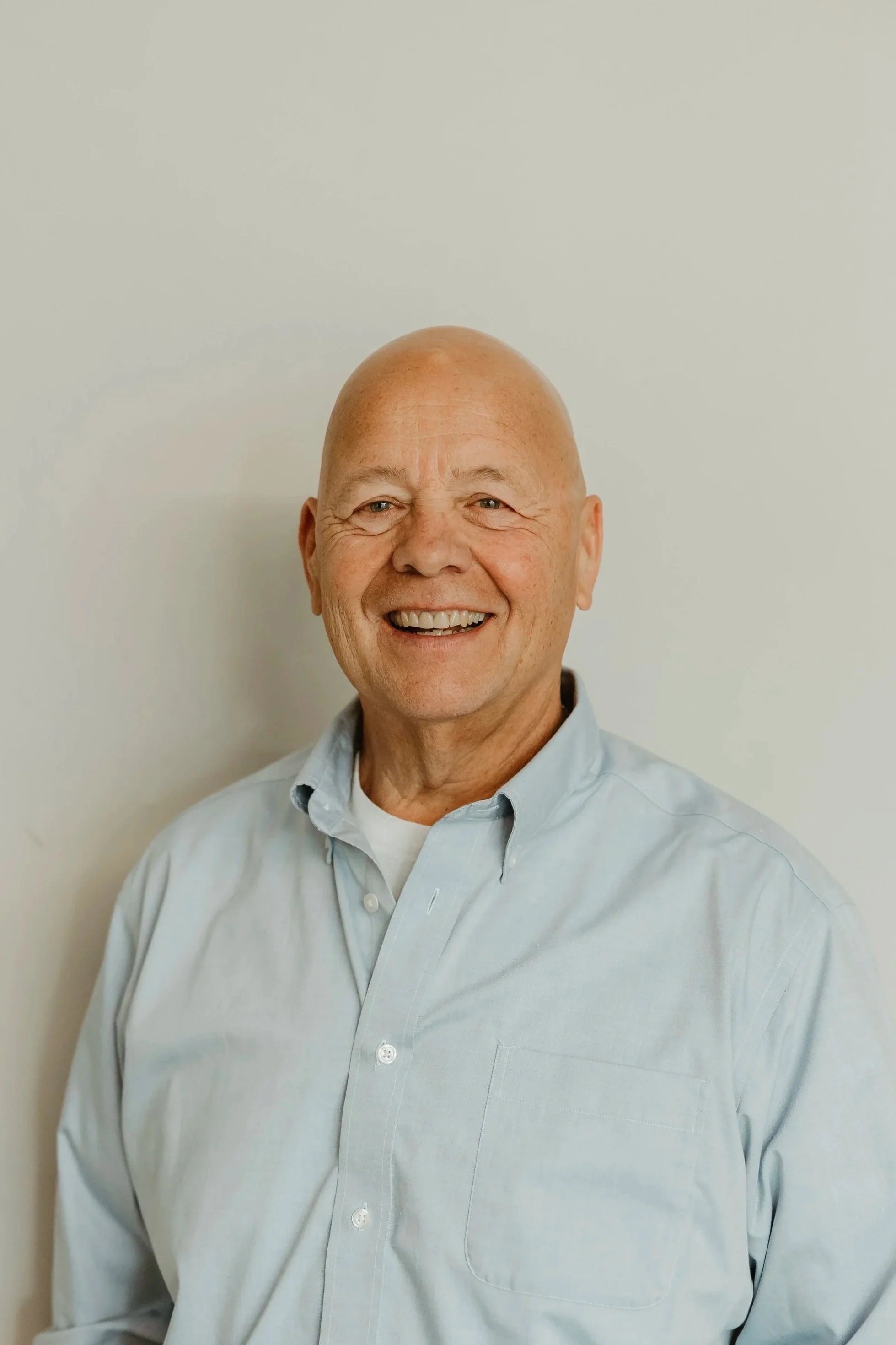 A smiling man wearing a light blue button-up shirt against a plain light background.