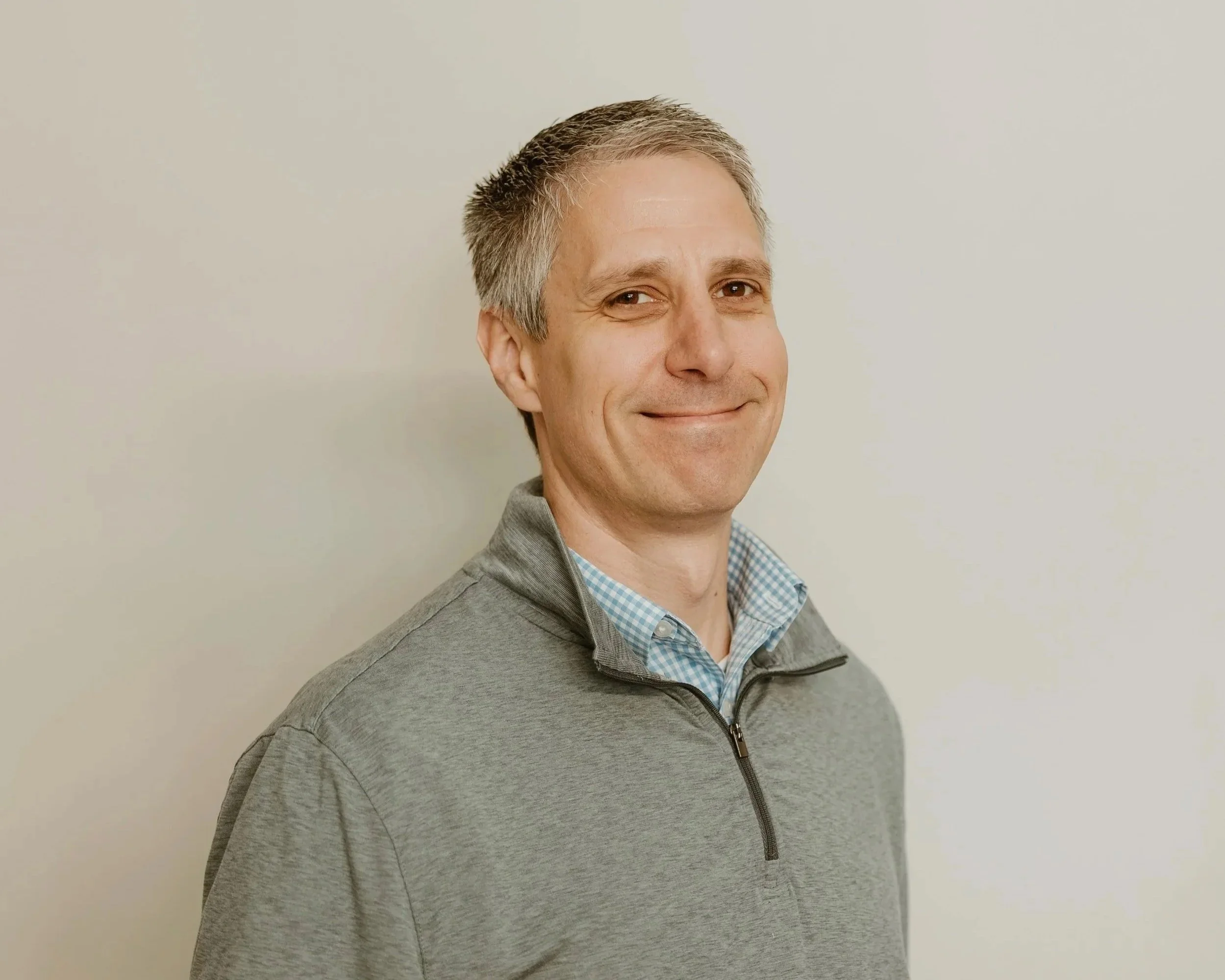 Headshot of a smiling middle-aged man with short, wearing a gray zip-up jacket over a blue checkered shirt, against a plain off-white wall background.