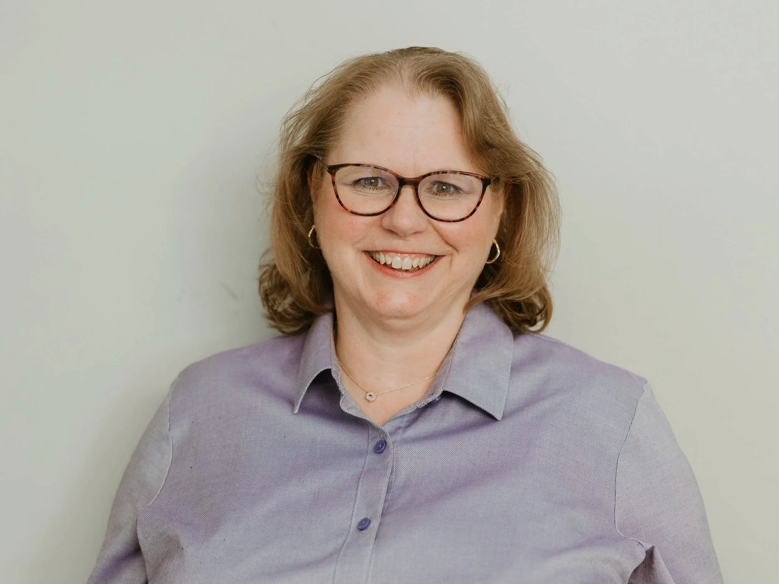 A woman with red hair, glasses, wearing a lavender blouse, smiling with visible teeth, in front of a plain white wall.