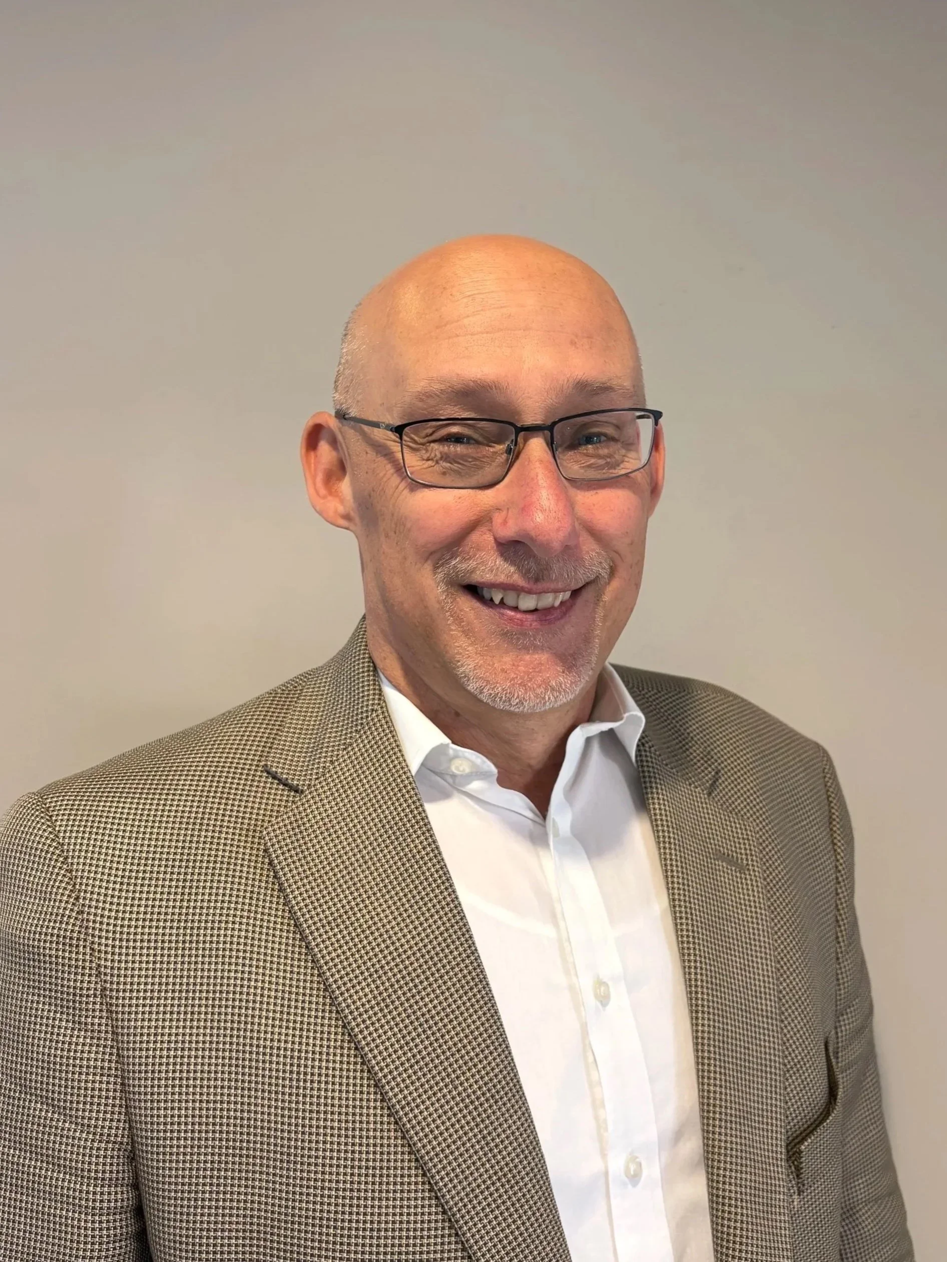 A middle-aged man with glasses, and a goatee, smiling. He is wearing a brown checked blazer and a white shirt, standing against a plain light gray background.